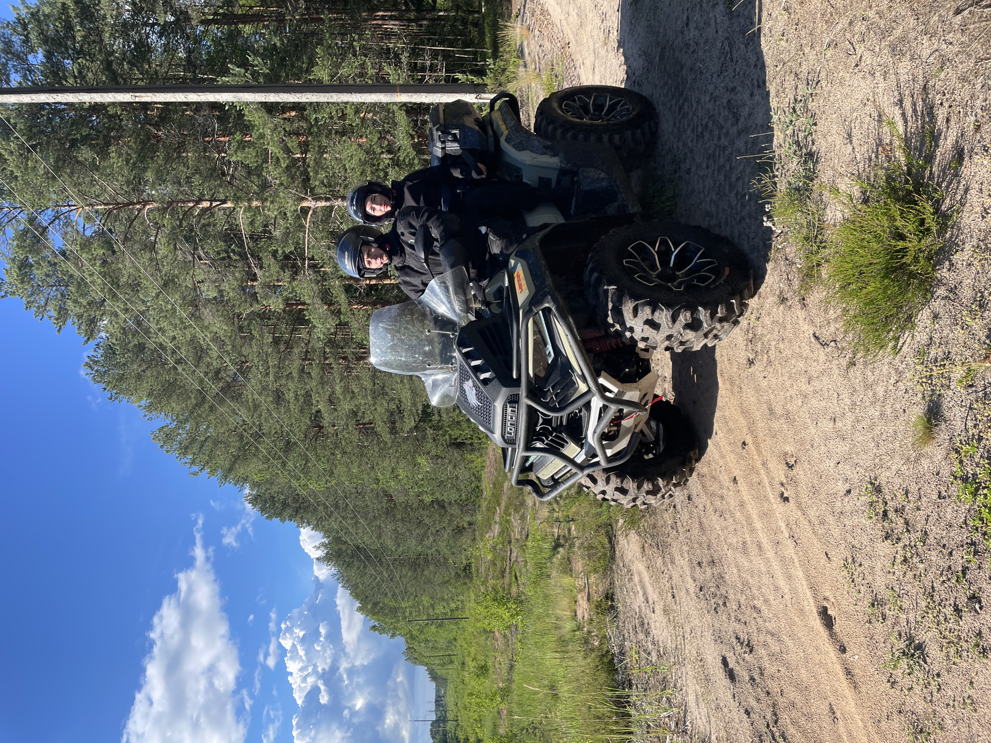 Two individuals, a male driver and a female passenger, are seated on a green and black all-terrain vehicle (ATV) on a sandy dirt road. Both are wearing helmets and black riding gear. The ATV has large, knobby tires and a windshield. The scene is set in a wooded area with tall pine trees and green undergrowth. The sky is blue with scattered white clouds. The location is identified as Bol'shie Ozerki, Russia.