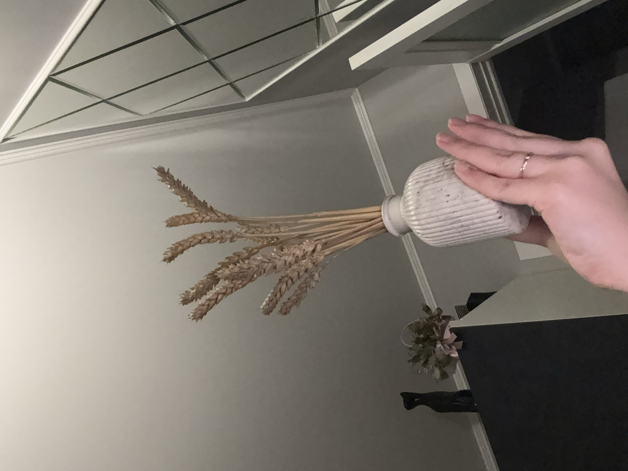 The image, when correctly oriented, captures an indoor scene primarily featuring a human hand holding a vase filled with dried wheat stalks. A fair-skinned hand, wearing a subtle ring on the fourth finger, firmly grips a light-colored, vertically ribbed ceramic vase or bottle. From the vase, numerous dried wheat stalks with prominent, golden-brown ears fan out towards the upper left of the frame.
The background reveals an interior setting. A distinct grid-patterned ceiling, possibly comprising square or rectangular panels with visible framing, occupies the upper portion of the image. Below the ceiling, a light-colored wall with architectural molding or trim is visible. Towards the lower right, on a dark, flat surface—likely a table or shelf—rests a small decorative item, resembling a faux plant arrangement with light foliage and a touch of pink. Beside this, a dark, elongated figurine or object, possibly a stylized cat, stands. The lighting is subdued, suggesting an indoor environment without strong natural light, or perhaps an evening setting. No text is visible in the image.