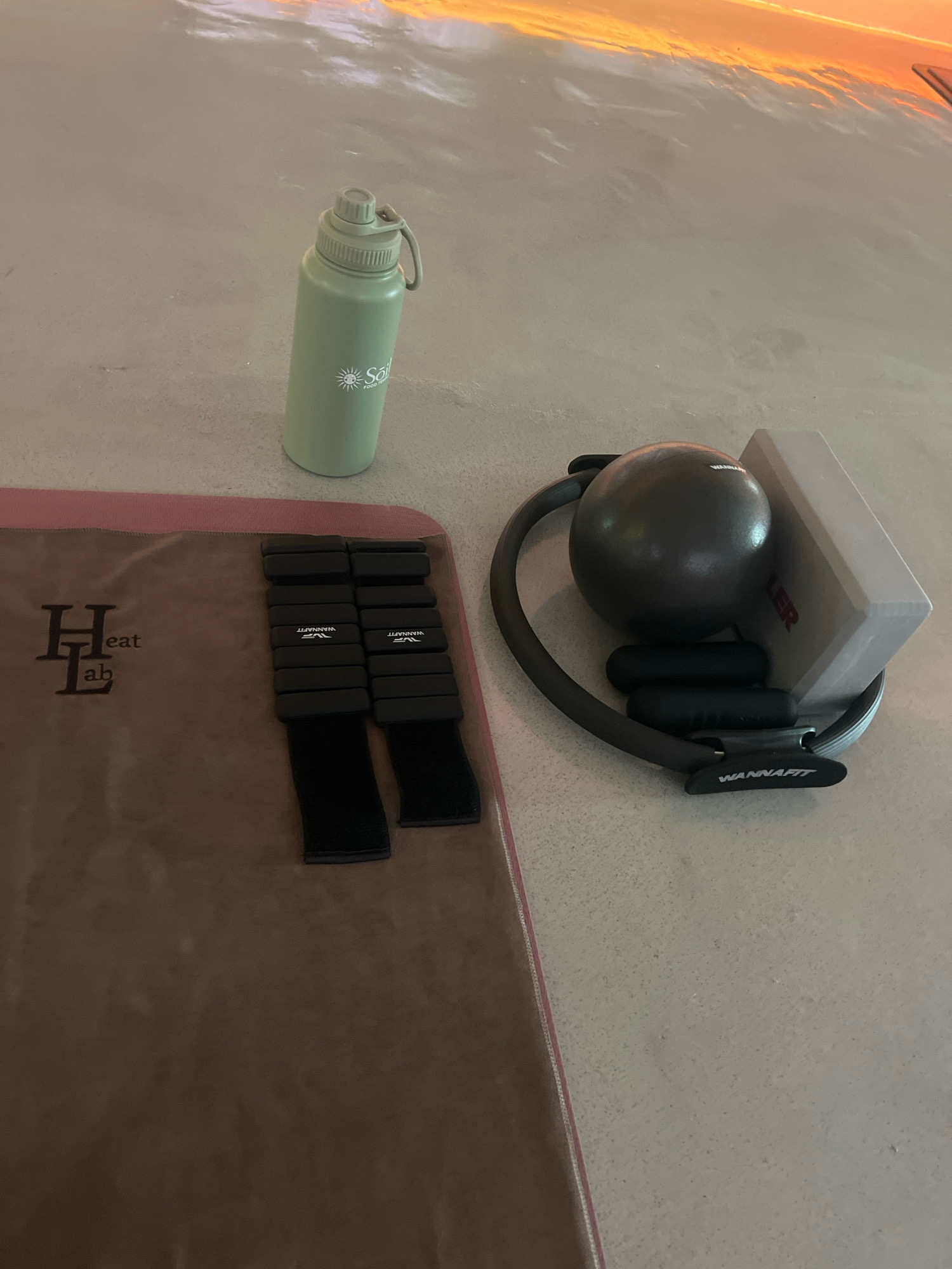 In North Kuta, Indonesia, a fitness setup is arranged on a textured gray surface. A light green water bottle with a white sun logo is positioned to the left. To the right, a collection of exercise equipment is grouped: a dark gray Pilates ring, a small black exercise ball, two black handles, and a light gray rectangular yoga block. In the foreground, a dark brown exercise mat is visible, featuring a stylized "HL" logo with the word "eat" and "ab" underneath. Two sets of black ankle weights are placed on the mat, each consisting of several weighted segments. The background shows a reflective surface with an orange and yellow gradient, suggesting it might be a window or a polished floor reflecting an sunset. No people or actions are depicted.