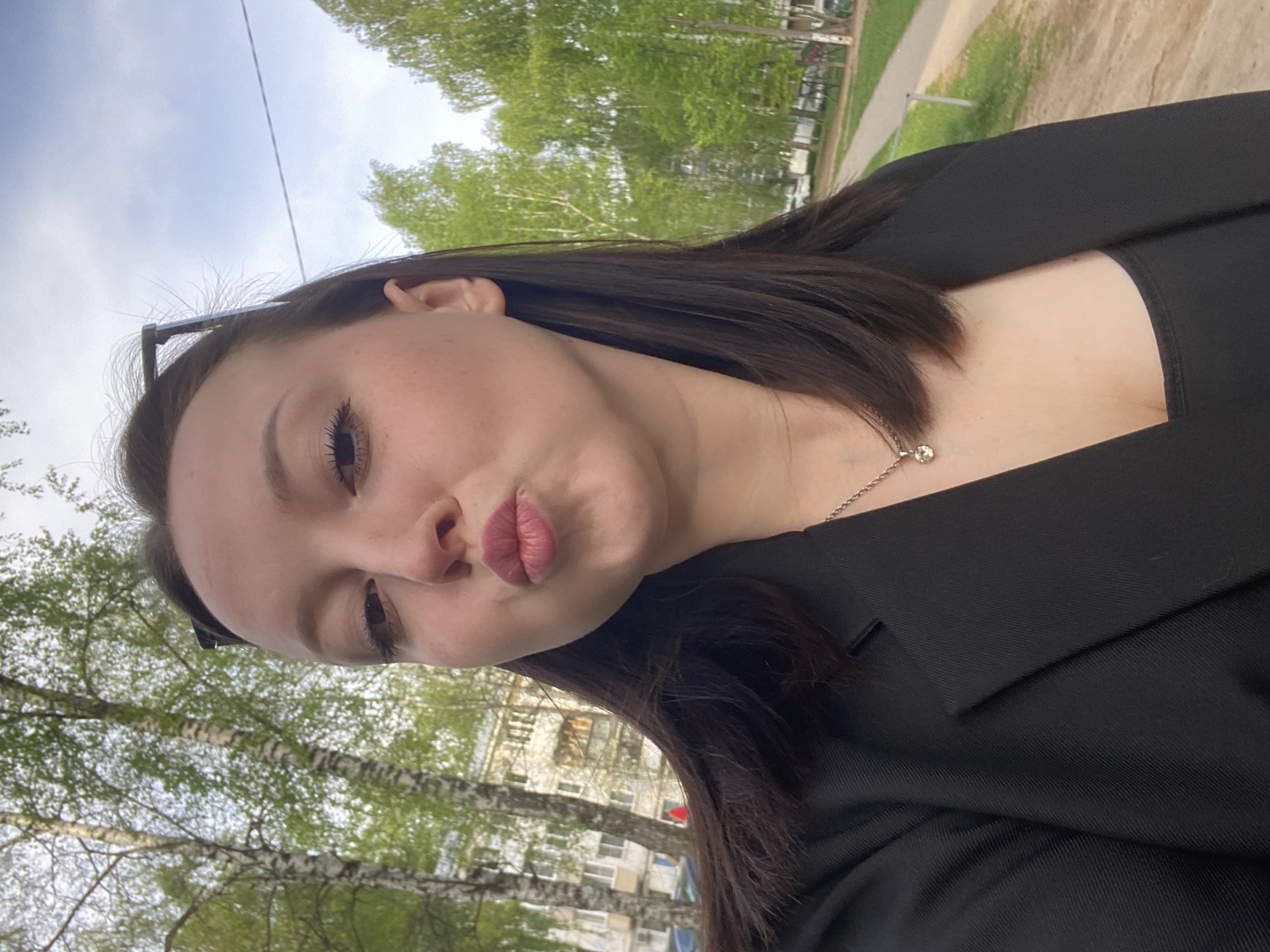 A young woman with dark hair and fair skin is taking a selfie outdoors. She is wearing a black blazer and a delicate silver necklace. She has her lips pursed in a kissing expression. The background shows trees with green foliage, likely birch trees based on their white bark, and a building with multiple windows, suggesting an urban environment. This image was taken outdoors in the city of Chaikovsky, Russia.