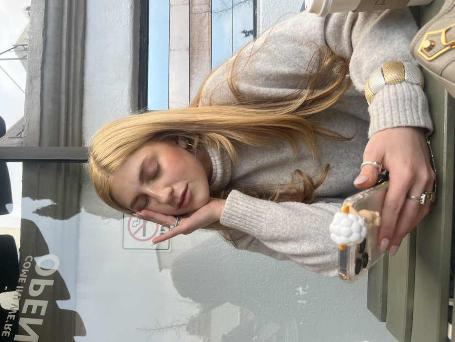 A young woman with long blonde hair and a light-colored sweater rests her head on her hand, her eyes closed. She is wearing a gold and silver bracelet and several rings. A clear phone case with a cloud-like decoration sits in her hand. An "OPEN" sign and a "no smoking" symbol are visible in the background, as well as part of a window and a coffee cup.