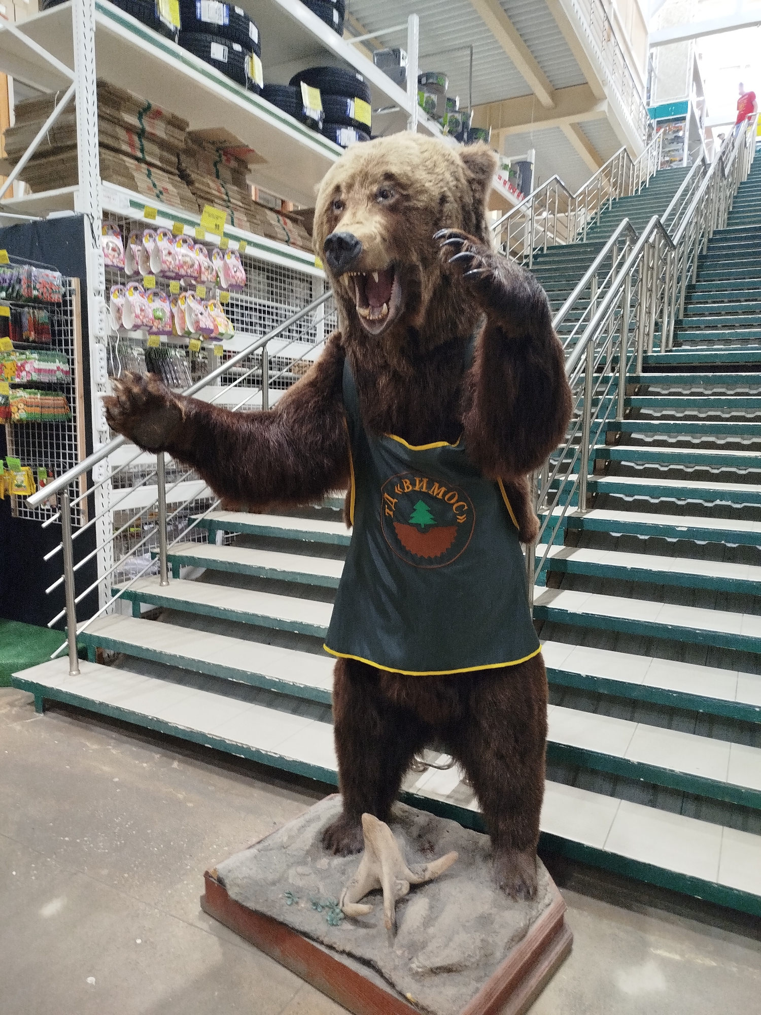 An artificial bear, dressed in a dark green apron with a circular emblem featuring a green tree and brown background, stands on a stone base with a wooden trim. The bear is positioned in an indoor setting, likely a retail store, with a set of metal and green-painted stairs behind it. Shelving units filled with various products, including tires and packaged goods, are visible in the background. The floor appears to be concrete. A person in a red shirt is visible on the upper stairs in the distance. The overall scene suggests a display within a commercial establishment.