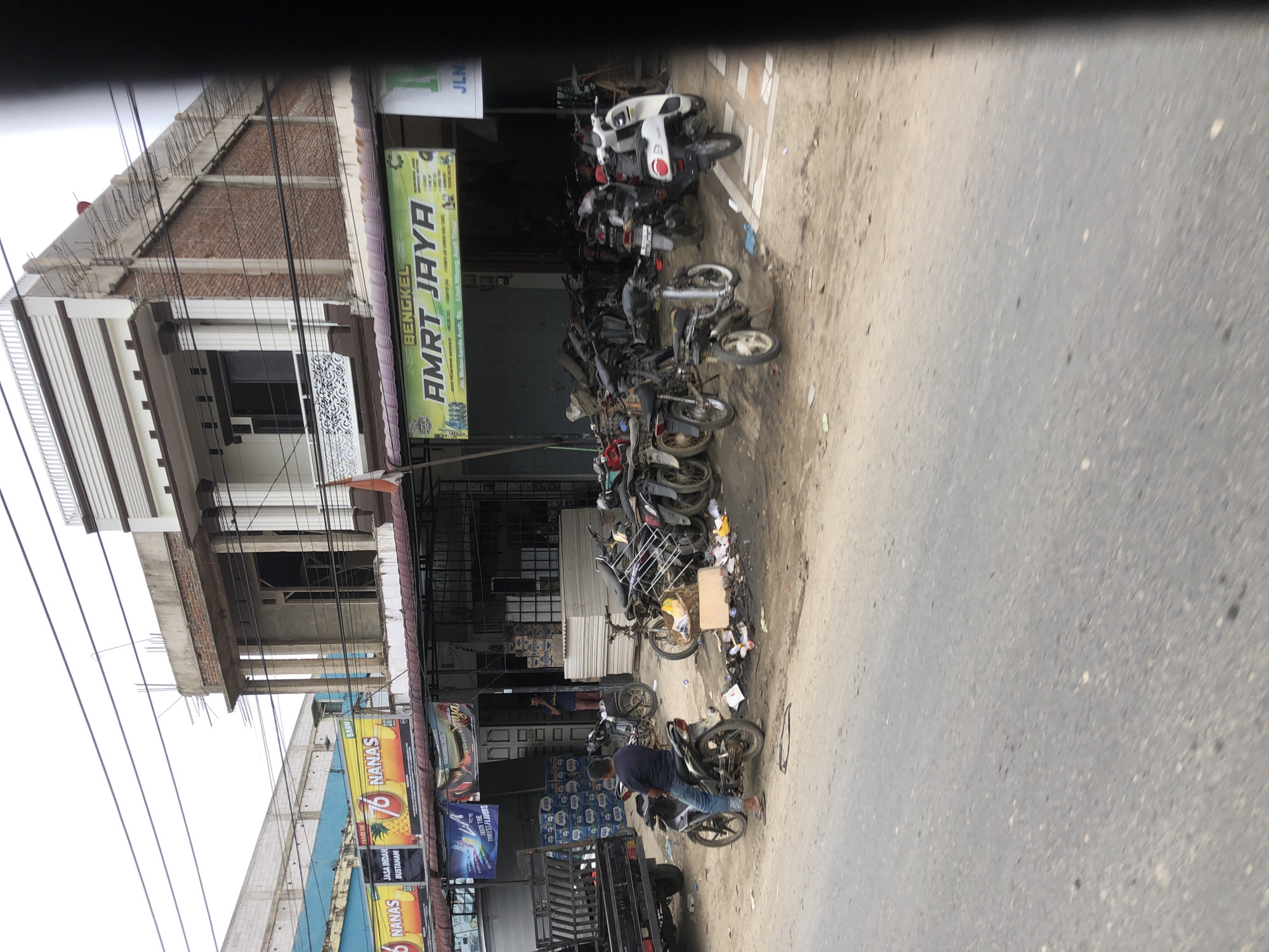 The image shows a street scene in Keudebaro, Indonesia. On the left is a partially constructed multi-story building. Adjacent to it is a row of shops or businesses. One of these businesses is "BENGKEL AMRT JAYA" a repair shop located at Jin. Medan-Banda Aceh, No Lihok Nibong, Aceh Timur. The street is lined with parked motorcycles, some of which appear to be under repair or disassembled. A person is on one of the motorcycles. There is also a pick up truck in front of the row of shops or businesses. A billboard for "NANAS" a fruit juice product can be seen as well. The general environment appears somewhat cluttered and dusty.