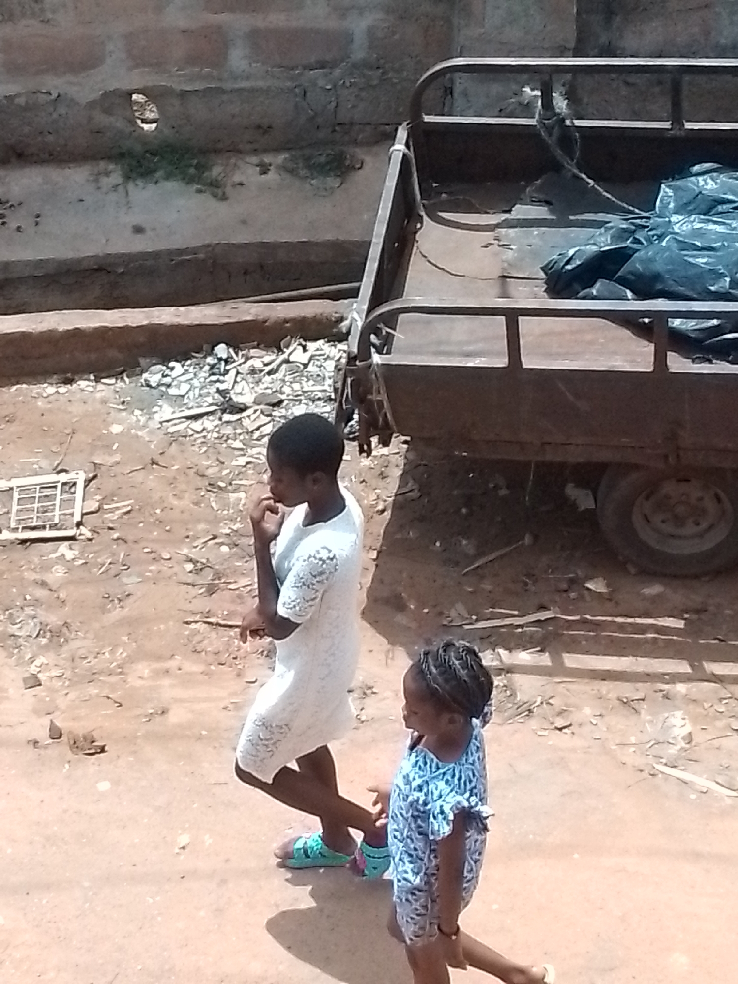Two people walk on a dirt street, in the city of Unknown, Unknown. The first person, an adult, is wearing a white dress and green sandals. The second person, a child, is wearing a blue and white patterned dress. To the right of the people is a trailer with a black tarp covering something in the back. The ground around them is littered with debris.