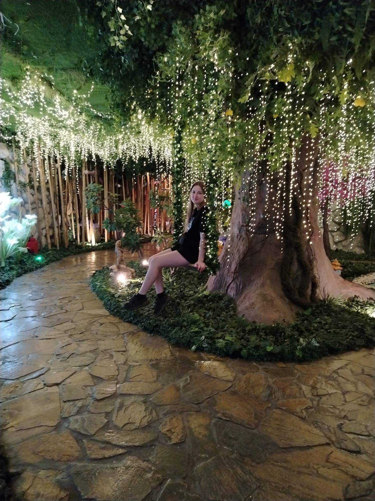 A woman is sitting on a tree branch in an indoor environment illuminated with strings of lights, likely an event space or restaurant. The floor is paved with irregularly shaped stone tiles. The background features artificial trees and foliage, with some areas covered in dense green plant life. The location is in the city of Kazan, Russia.