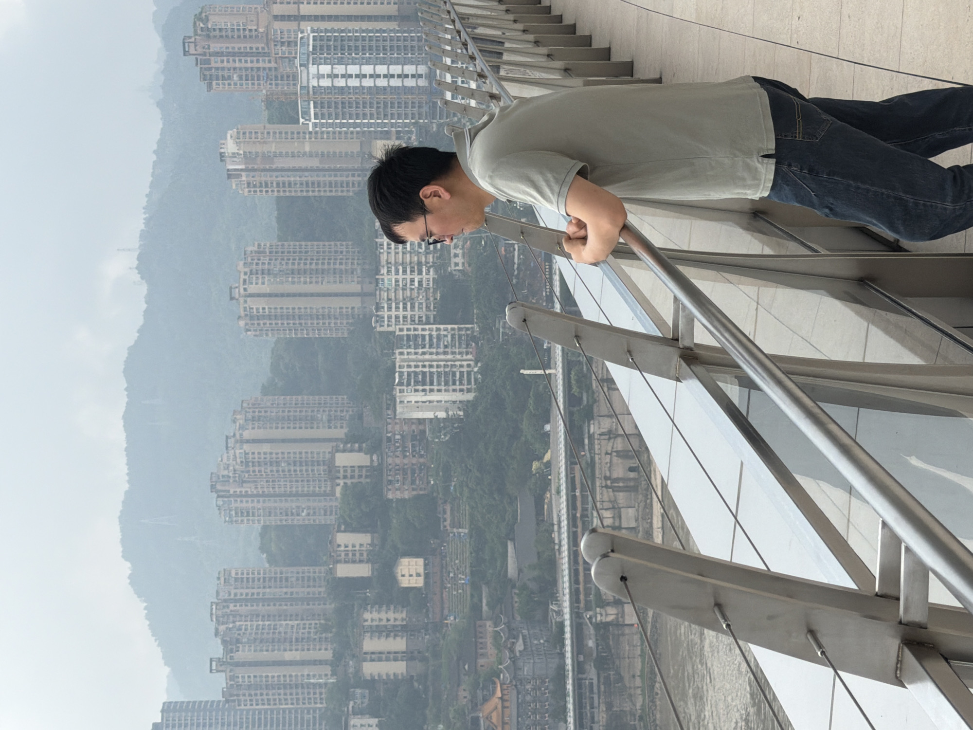 A man stands on a pedestrian walkway, leaning his arms on a railing and looking out at a cityscape. The walkway has a modern design with metal railings and glass panels. In the background, a densely populated city rises on a hillside, characterized by numerous apartment buildings and some older structures. A river runs through the city, with bridges spanning it. The sky is overcast, contributing to a hazy atmosphere. The man is wearing a light-colored polo shirt and dark jeans. The location is the city of Yuzhong District, China.