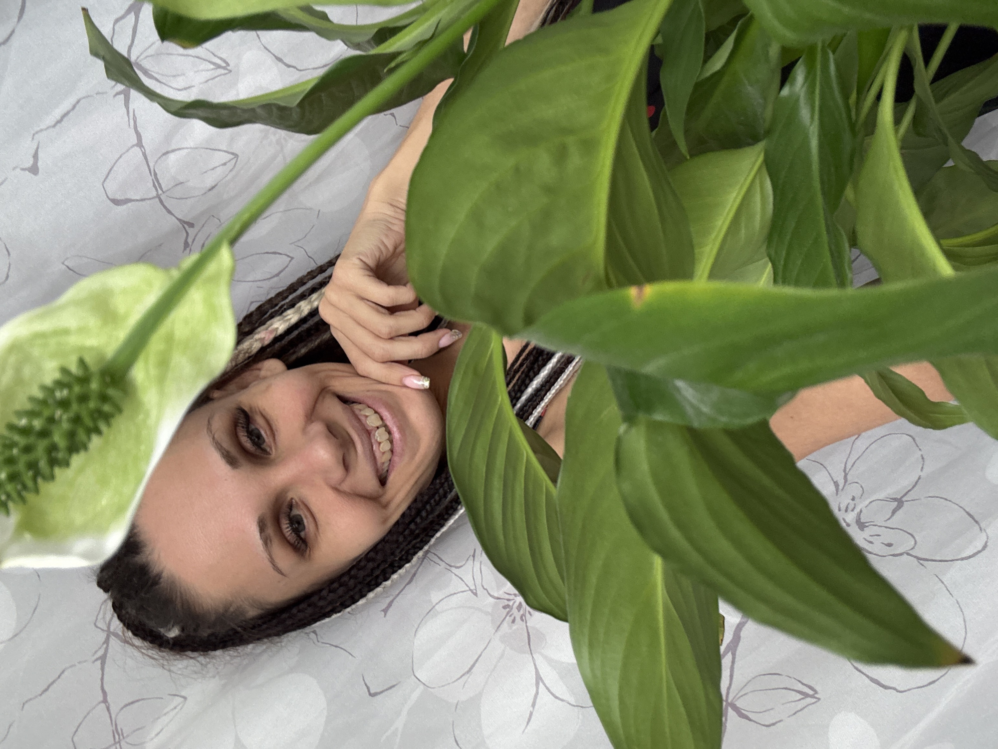 The image shows a smiling woman partially framed by the large green leaves of a plant, viewed from an overhead perspective. The woman is lying on a bed covered with white bedding that features a light grey floral pattern. Her head is positioned towards the upper left of the frame, tilted on her back, with her body mostly obscured by the foliage. She has dark, finely braided hair and is looking directly at the camera with a wide, cheerful smile, revealing her teeth. One of her hands is visible near her face, showing manicured fingernails with clear polish and subtle adornment.
Dominating the foreground and much of the right side of the image is a lush green plant with prominent, glossy leaves. A distinctive white spathe, characteristic of a peace lily, is visible in the upper left corner, partially out of focus. The plant creates a soft, natural frame around the woman's face, conveying a sense of intimacy and tranquility. The setting appears to be indoors, likely a bedroom, characterized by soft, ambient lighting. There is no visible text in the image. The woman's expression suggests happiness or contentment.