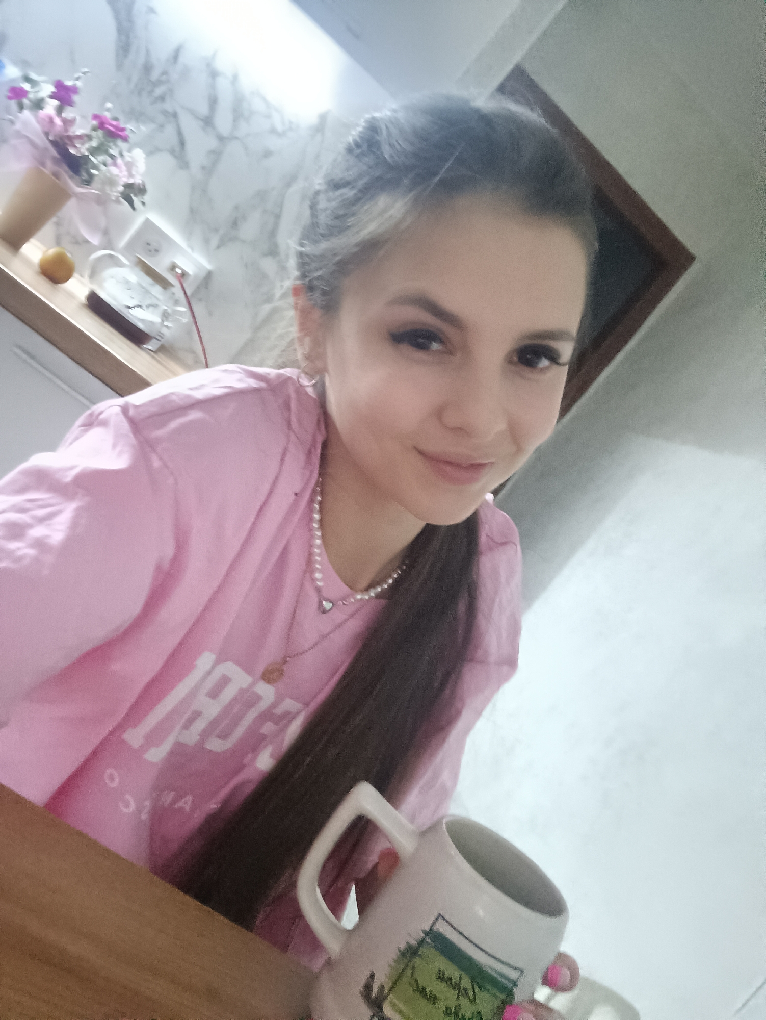 A young woman is seated at a table, holding a white mug with a green design, in what appears to be a kitchen or dining area. She is wearing a light pink t-shirt, a pearl necklace, and has dark hair styled in a low ponytail. The background includes a countertop with a floral arrangement, a clock, and a marble-patterned backsplash. The location is in the city of Unknown, Unknown.