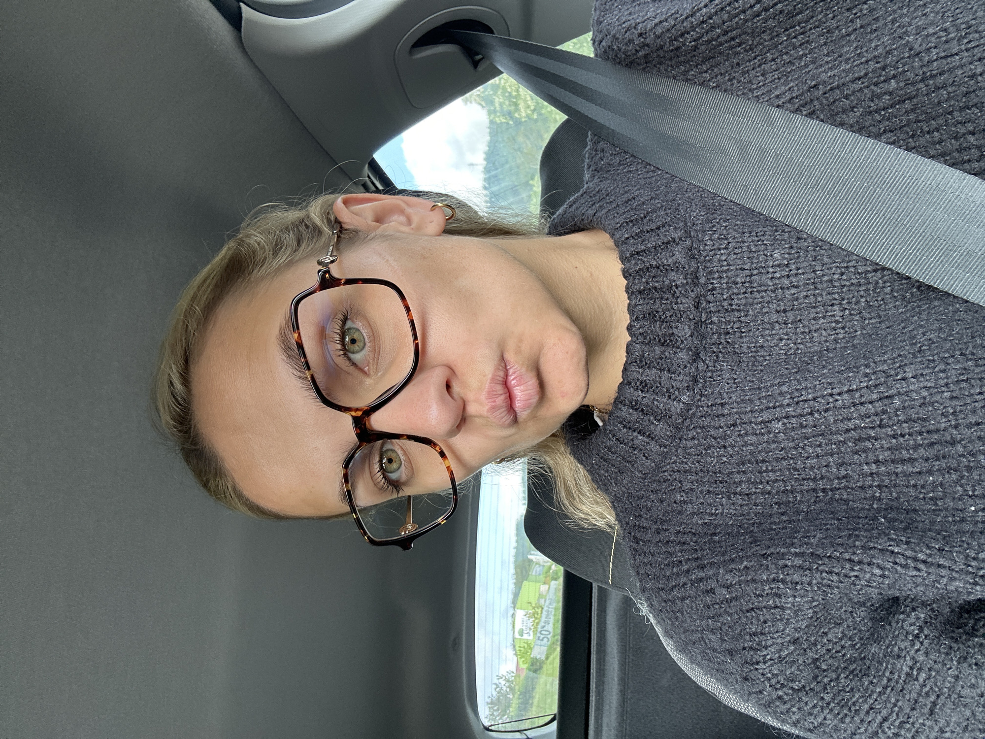 A medium close-up, portrait-oriented photograph captures a person with blonde hair and fair skin, wearing a dark grey knit sweater and tortoiseshell-framed glasses, seated in what appears to be the passenger seat of a car. The image is rotated clockwise, so the person's head is tilted significantly to the right, almost horizontal in the frame. They look towards the camera with a slight pout on their lips, their green eyes visible through the glasses. A dark grey seatbelt is fastened across their chest, running diagonally from the top right to the bottom left of the frame. Through the car window, a bright, clear daytime sky and lush green trees are visible, along with distant structures and a partially obscured sign displaying the number '150'. The scene depicts a casual moment inside a vehicle, likely during the day, in Lorette, France.