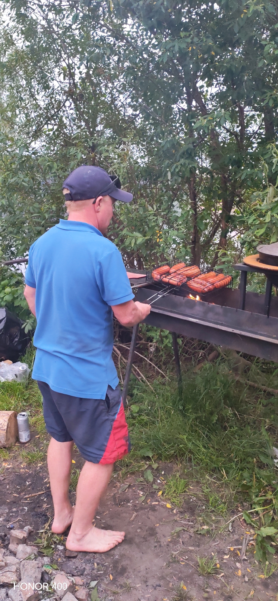 A man wearing a blue polo shirt, dark shorts with red trim, and a dark blue baseball cap is standing barefoot on dirt and grass, grilling sausages over a fire in what appears to be an outdoor, possibly rural setting. He is holding a grill basket with several sausages in it over a black metal barbecue. The background consists of dense green foliage, suggesting a natural environment. The image is a vertical shot, with the man positioned in the center left. There is a can and some firewood visible on the ground to his left. The overall impression is a casual, outdoor cooking scene. The location is not specified in the image.