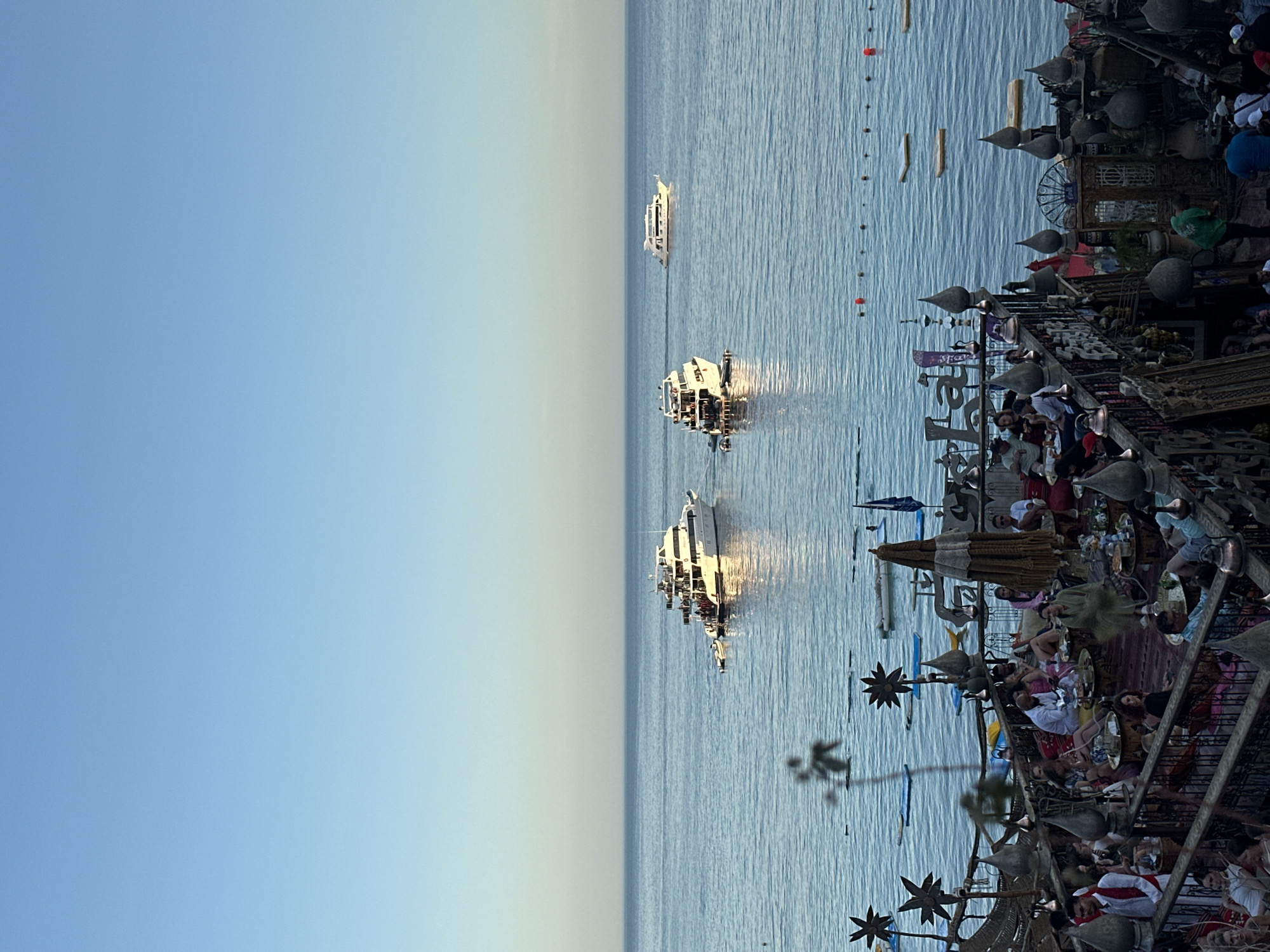 The image captures a vibrant waterfront scene during what appears to be late afternoon or early evening, with a clear sky transitioning from light blue to a pale, warm hue near the horizon.
In the foreground, on the right side of the image, a bustling promenade, pier, or restaurant area is visible. It features an elaborately decorated structure with intricate metalwork, ornate finials, star-shaped embellishments, and decorative screens, suggesting a traditional Middle Eastern or Moorish architectural style. Numerous people are present, seated at tables adorned with drinks and food, engaging in conversation and dining. Cushions, large umbrellas, and decorative elements like urns or large lamps contribute to the lively atmosphere. The area is well-lit, possibly by artificial lights within the structure and the natural light from the setting sun.
The midground is dominated by calm, deep blue water, reflecting the bright sunlight in dazzling streaks. Several boats and yachts are scattered across the water. The closest large vessel, a white yacht, is brilliantly illuminated by the sun's reflection, with passengers visible on its deck. Further out, two other white boats are also caught in the sun's glow. A line of small, dark buoys or markers, interspersed with a couple of red ones, extends into the water from the right edge. Smaller watercraft, possibly paddleboards or dinghies, are also seen closer to the shore.
The background consists of a clear, expansive sky, forming a gentle gradient from a deeper blue on the left to a lighter, almost white-blue towards the right horizon, where it meets the water. The overall impression is one of leisure and enjoyment in a picturesque seaside setting. There is no visible text in the image.