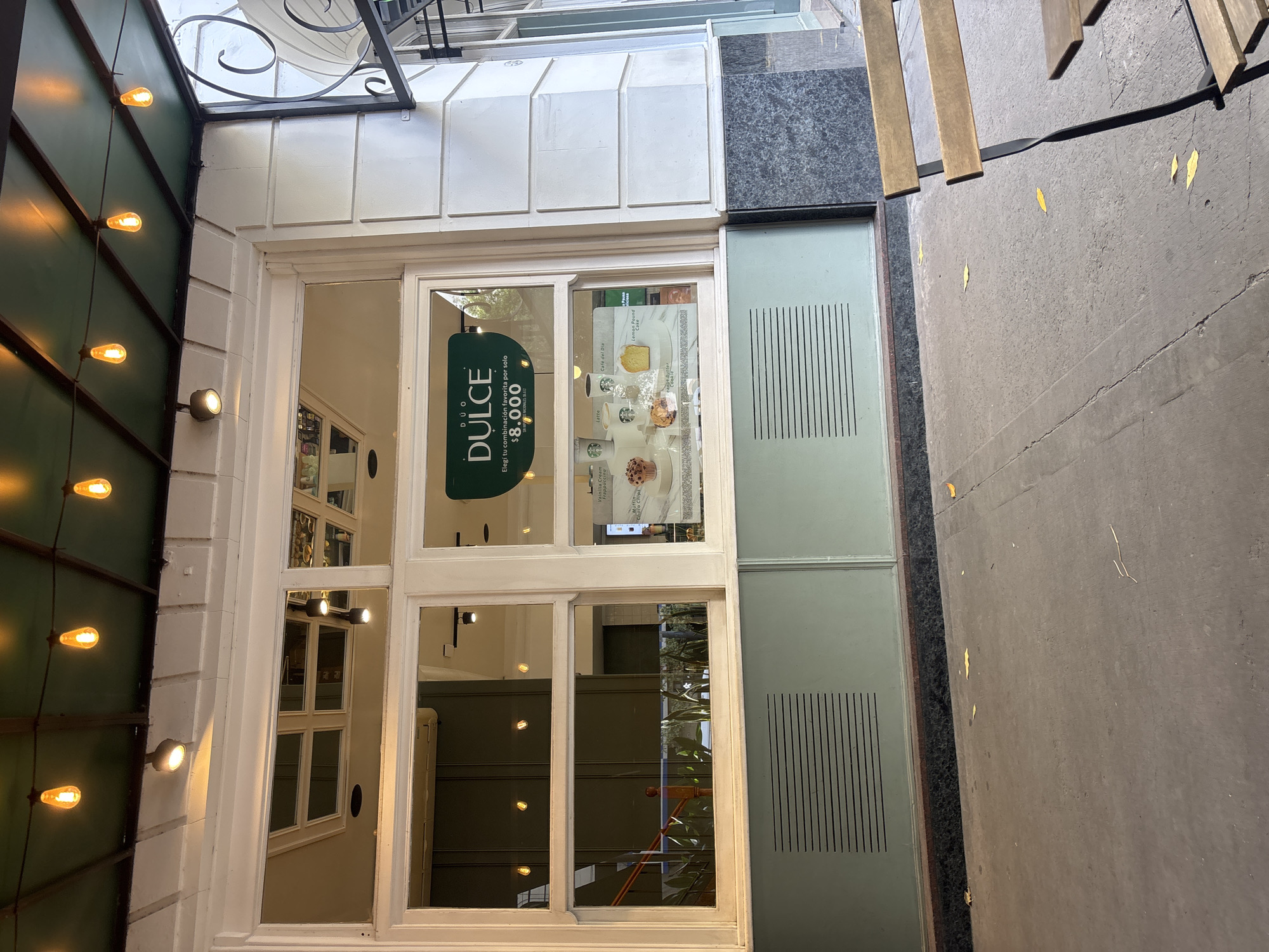 The image shows a street view of a business in Buenos Aires, Argentina. The business, called "Dúo Dulce", is visible through a window that is divided into multiple panes. A green sign bearing the business name "Dúo Dulce" and the price "$8,000" is prominently displayed. Inside the window, menu items like "Latte," "Café del Día," and "Lemon Pound Cake" are advertised.
The exterior of the building has a white facade with decorative molding and a green wall adorned with string lights. Next to the window, there's a gray panel with a vertical line pattern. The sidewalk is gray with scattered leaves and a wooden bench is partially visible. The perspective of the image is somewhat tilted.