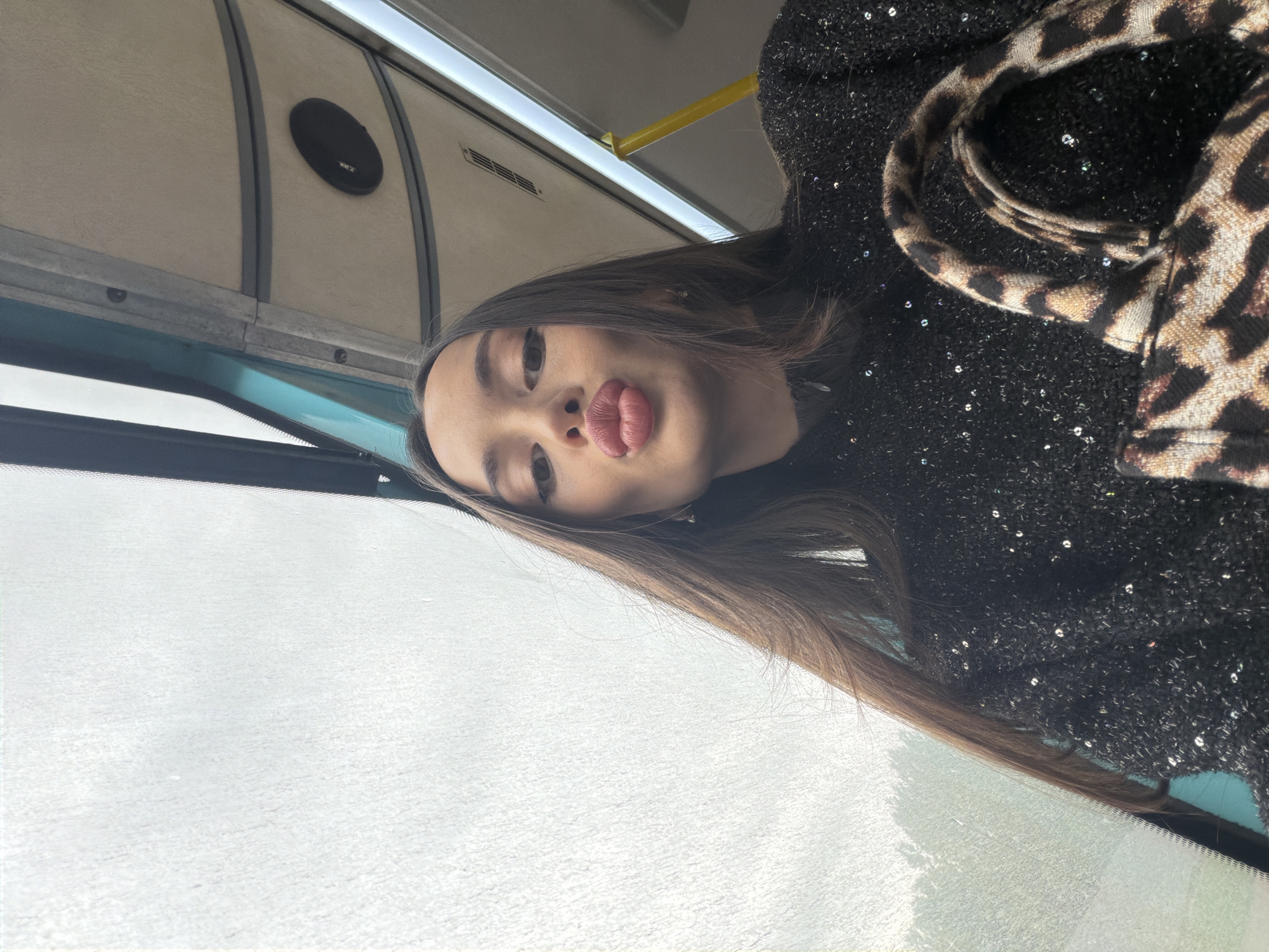 A woman is in a vehicle, possibly a bus or train, in what could be Saint Petersburg, Russia, based on the architectural details visible through the window. The interior of the vehicle features overhead panels with a speaker and structural elements. The woman is wearing a black sparkly garment and a leopard-print item. The woman is positioned in front of a window, possibly looking out. She is wearing makeup and making a "duck-face." The background features a blurry urban landscape.