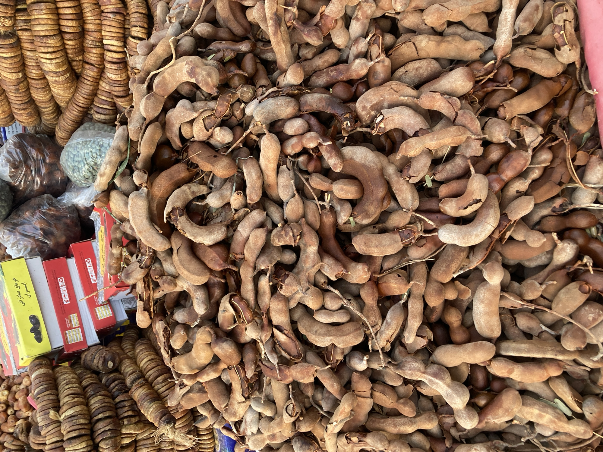 A high-angle shot shows a large pile of tamarind pods dominating the scene. The pods are brown and varied in size and shape. To the left of the tamarind, there are strings of dried figs stacked in rows. Also visible are boxes with Arabic script, possibly product packaging, and plastic bags containing what appears to be seeds or nuts. The overall composition suggests a marketplace setting, likely in Quetta City Tehsil, Pakistan, where such items are commonly sold.