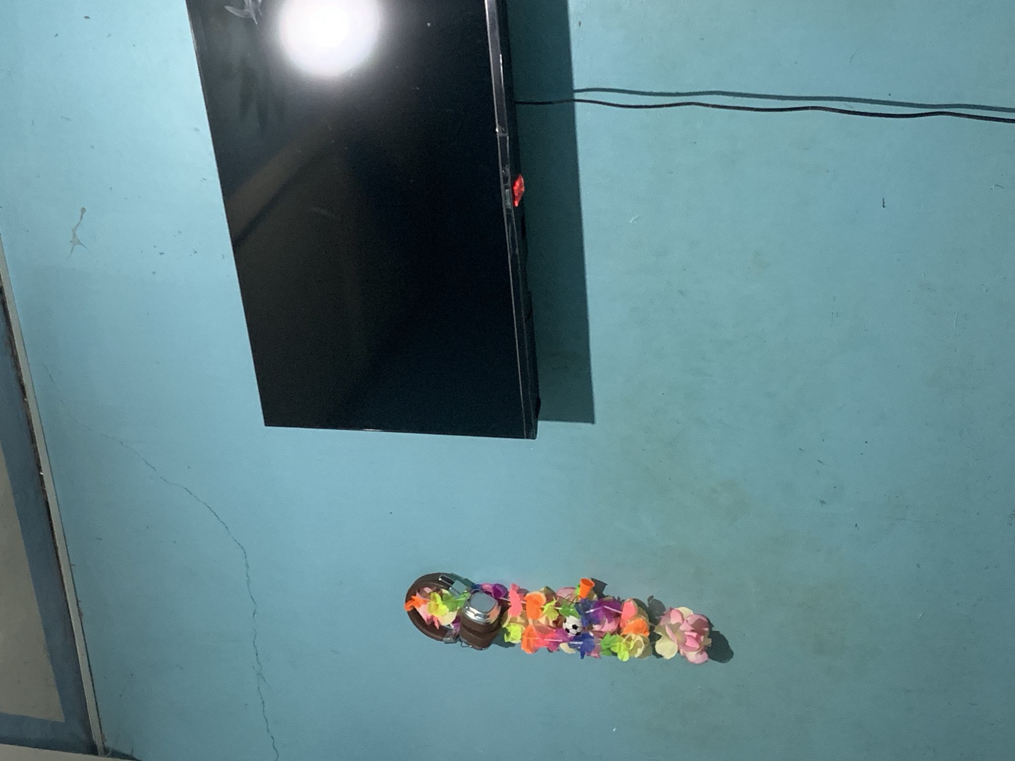 A framed television is mounted on a light blue wall in a room. To the left of the television, a string of colorful artificial flowers hangs vertically, with a pair of headphones at the top. A thin black cord runs vertically down the wall to the right of the television. The wall shows signs of wear, with cracks and some dirt marks. The lighting suggests an indoor environment. The location is a room in Santa Ana, Venezuela.