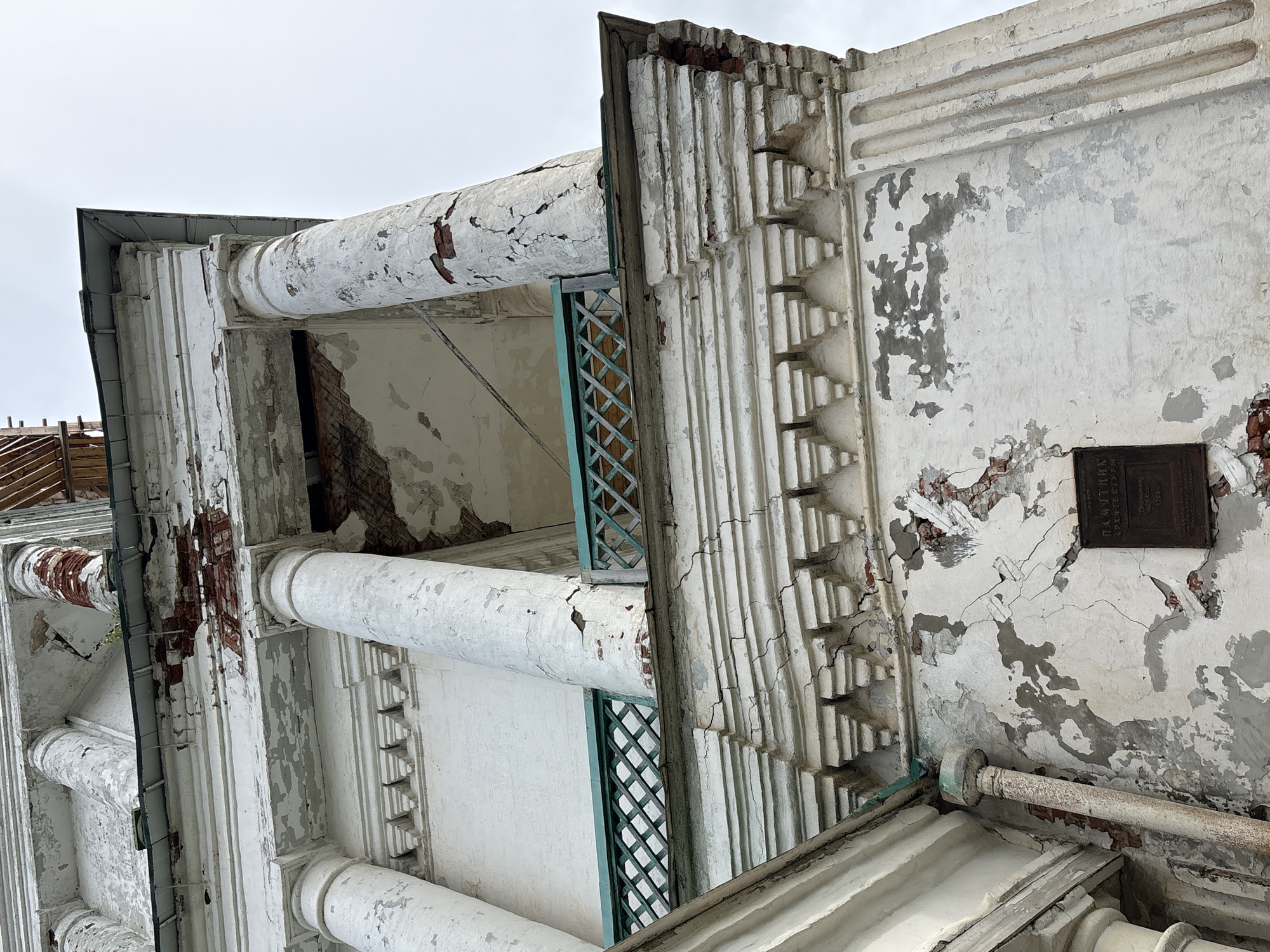This is a close-up, low-angle, exterior shot of the weathered bell tower of the Church of the Resurrection in Kstynino, Russia. The bell tower, constructed primarily of white brick, exhibits significant deterioration. Large sections of the white plaster are peeling away, revealing the red brick beneath. Cracks are visible in the plasterwork of the columns and on the main body of the tower. A green, lattice-like railing runs along a balcony or walkway. A dark metal plaque is affixed to the wall, bearing Cyrillic text. The sky is overcast and appears white. The image focuses on the architectural details and the visible signs of decay.