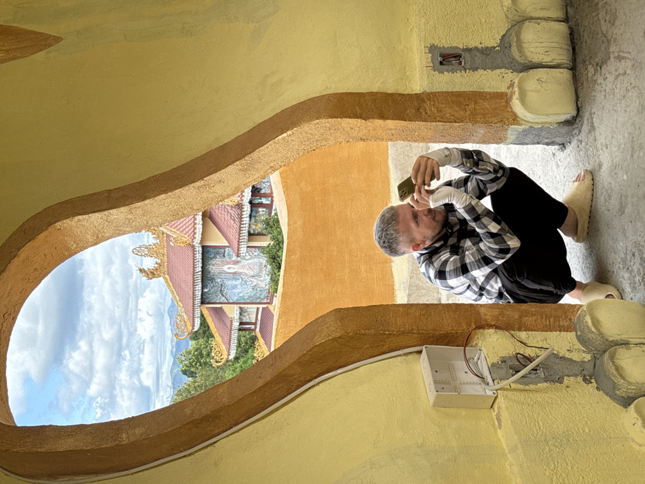 The image depicts a man crouching within a yellow-painted archway, observing a smartphone, with a traditional Vietnamese temple visible through the archway in Đăk Mar, Vietnam. The man, a Caucasian male with short grey hair, is wearing a black and white plaid shirt, black pants, and light-colored slip-on sandals. His forearms are covered with white wraps or sleeves. He holds a green-cased smartphone with both hands, focusing on its screen. The interior walls of the structure are painted a textured yellow. To the left, an open electrical box with exposed wires is affixed to the wall, and to the right, a similar conduit entry is visible.
Beyond the archway, an orange-painted wall extends across the midground. Above this wall, a multi-tiered building, characteristic of a Buddhist temple, is visible. It features intricate red-tiled roofs with decorative gold finials and ornamentation. The temple's facade displays a large mural depicting a white deity, likely Buddha, set against a background of stylized trees and foliage. Green trees are interspersed around the temple structure. In the distant background, rolling hills or mountains are silhouetted against a partially cloudy sky.
