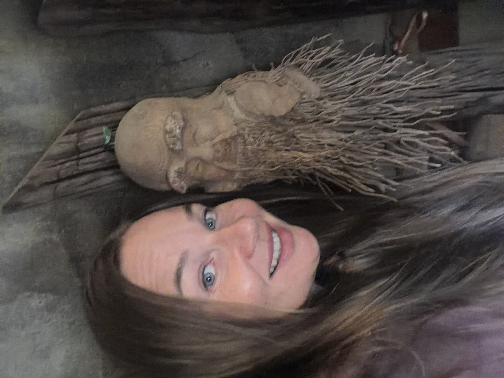 A selfie taken by a woman with long, brown hair and blue eyes, who is smiling at the camera. To her right is a carved wooden figure with a long, stringy beard and a small green leaf on its head. The background appears to be a rustic wooden structure. The image is taken indoors, likely in a place with natural or rustic decor. This location could be a shop or a display area within the city of Lâm Viên - Đà Lạt, Vietnam, known for its artisanal crafts and natural beauty.