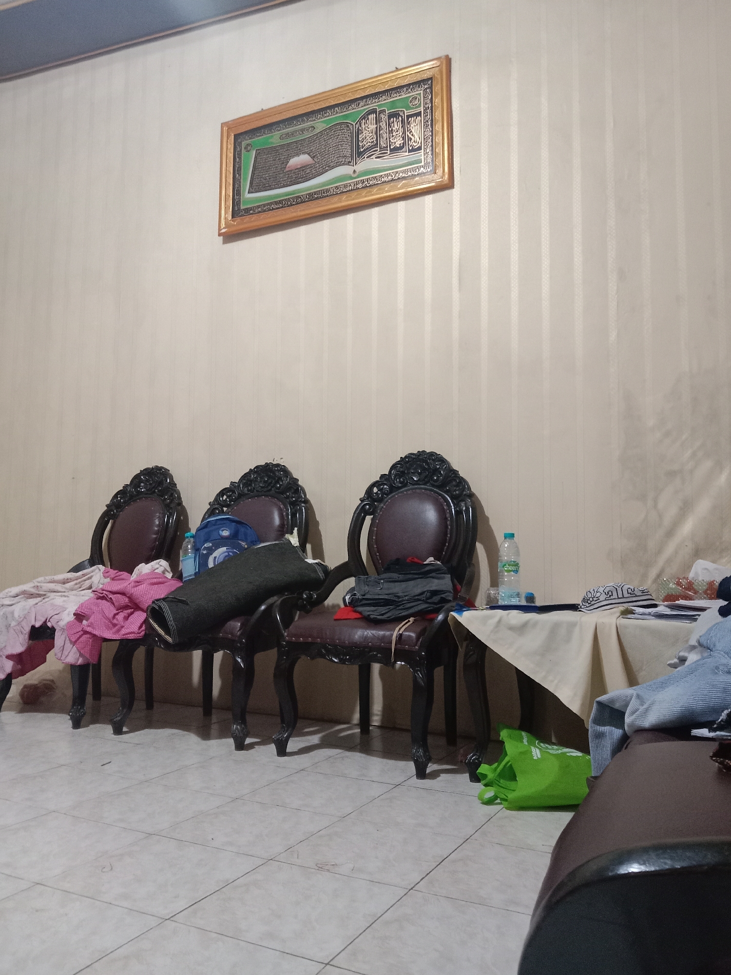 In a dimly lit room, possibly a living room or a waiting area in Madiun, Indonesia, three ornate, dark-wood chairs with plush, brown leather seats are arranged in a row. The chairs have intricate carvings on their backs and legs. The chair on the far left has a pink and white checkered cloth draped over its armrest and a dark, rolled-up blanket or mat lying across its seat. To its right, a blue backpack rests on the seat of the second chair, next to a partially obscured bottle of water. The third chair, on the right, has a stack of dark clothing on its seat.
Behind the chairs, a wall is covered in cream-colored wallpaper with a subtle vertical stripe pattern. A framed piece of art, featuring calligraphy and an image of what appears to be the Kaaba, is hung on the wall above the chairs.
In the foreground, a small portion of a brown sofa is visible on the right. To the left of the sofa, a light-colored tablecloth is spread over a surface, with a bottle of water and a white cap with intricate patterns on it. A green reusable shopping bag sits on the floor in front of the table. The floor is tiled with light-colored square tiles. The overall impression is of a somewhat cluttered, domestic interior.