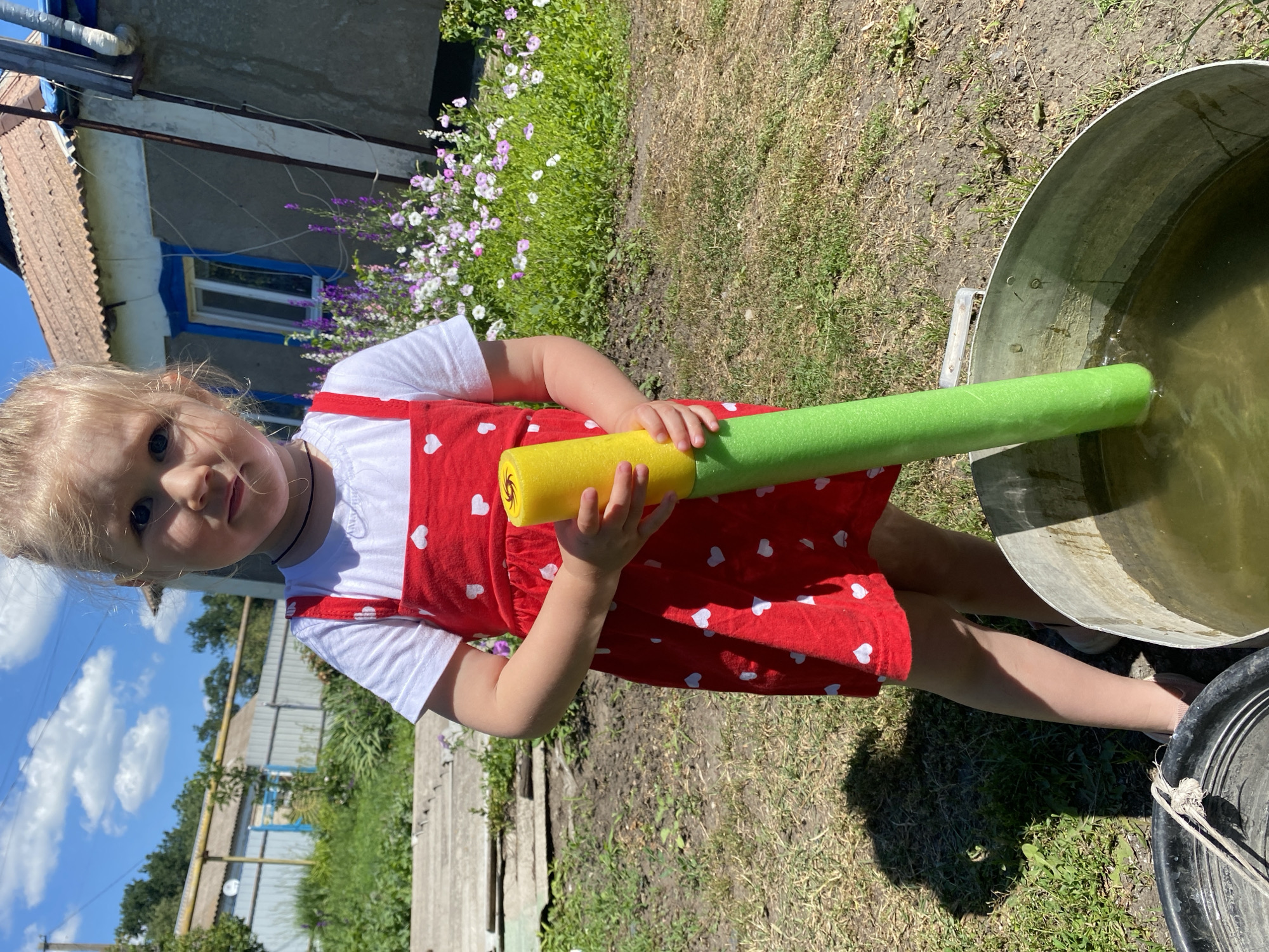 A young girl with blonde hair stands outdoors holding a yellow and green foam pool noodle. She is wearing a white t-shirt with a red pinafore dress adorned with white hearts. The girl is positioned in front of a house with a rustic appearance. The background features greenery, including a patch of purple and white flowers, and a bright blue sky with scattered white clouds. To the girl's right, a large metal tub filled with murky water is visible. A black plastic tub with a rope is to her left. The scene appears to be a residential area, possibly in the city of Mariupol, Ukraine, based on the context provided.