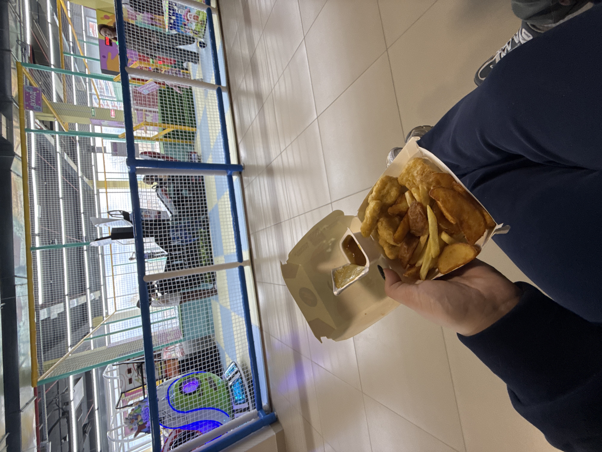 The image, rotated 90 degrees clockwise, captures an indoor scene in Perm, Russia, depicting a person holding food next to a large, multi-level children's play structure.
In the foreground, a person's hand holds a cardboard container filled with fried food, appearing to be a mix of french fries and possibly chicken nuggets or fish pieces. A small plastic cup of dipping sauce, likely honey mustard or similar, is also nestled within the container. The person is wearing dark blue pants and a dark blue long-sleeved garment, with a glimpse of sneakers visible on their feet. The floor is covered with light-colored square tiles.
To the left of the frame, occupying a significant portion of the background, is an elaborate indoor play area. This multi-story structure is constructed with metal frames, safety netting, and various colorful elements in yellow, blue, and green. Within the play structure, several platforms, ramps, and tubes are visible, creating a complex environment for children. In one of the cropped hints, a child's face can be seen peering out from an upper level of the structure. Another section shows what resembles a miniature basketball hoop.
Visible text within the play area includes a purple sign with the word "ВХОД" (VKHOD), which means "ENTRANCE" in Russian, indicating access to the structure. A yellow sign displaying the number "1" is also present, possibly marking a level or section. The overall lighting suggests a brightly lit indoor environment, possibly a family entertainment center, a shopping mall, or a similar leisure facility. The scene conveys a relaxed, casual atmosphere, typical of a family outing or a break during leisure activities.