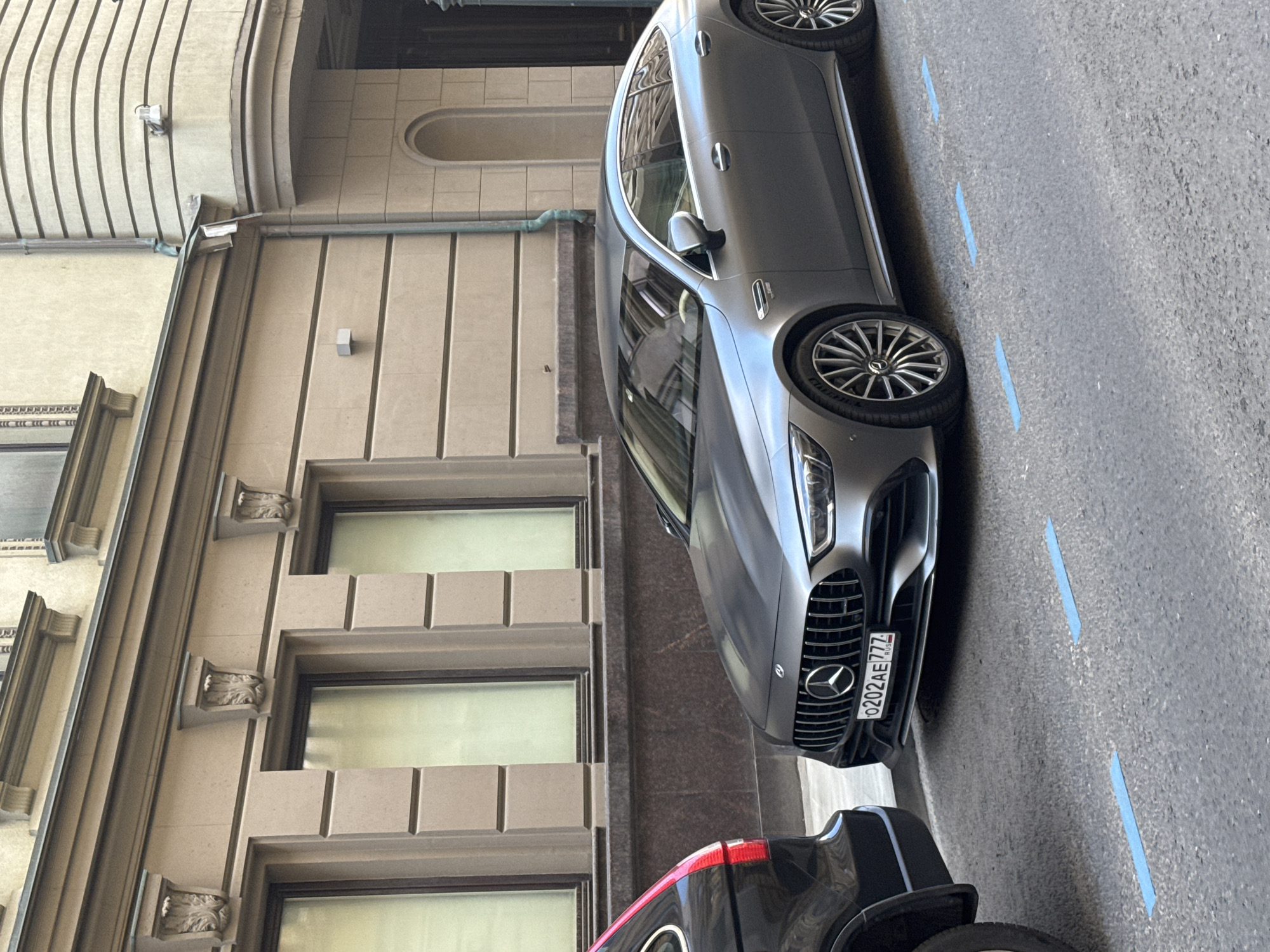 A silver Mercedes-Benz sedan is parked on a street next to a building with classical architectural features. The car has a license plate that reads "0202AE777 RUS". The building is made of light-colored stone and has several windows with decorative moldings. The street is paved with asphalt and marked with blue lines, possibly indicating parking spaces. The scene appears to be in an urban environment, likely Moscow, Russia, given the license plate. The lighting suggests it is daytime, and the weather appears to be clear. There are no people visible in the image.