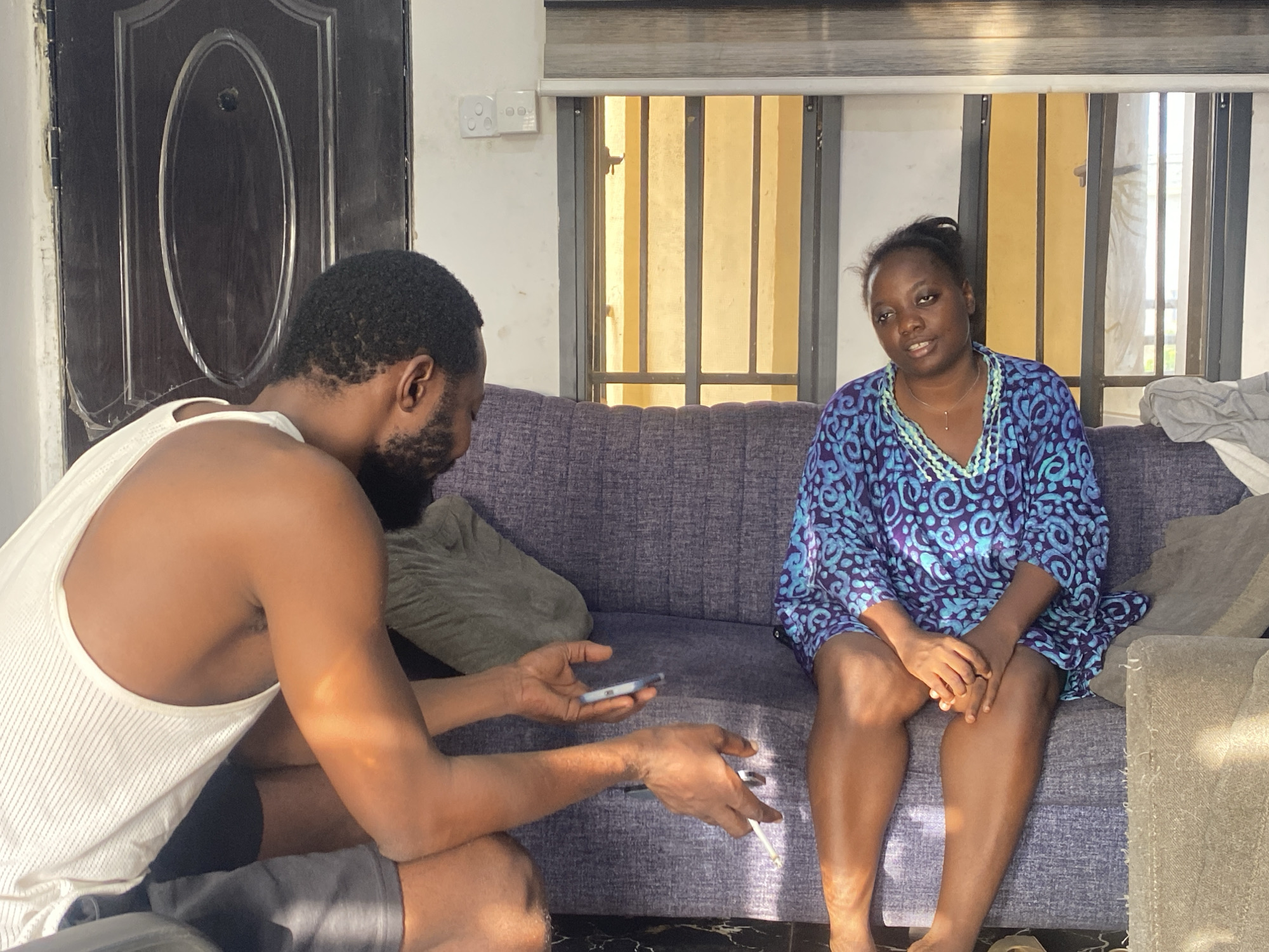 This indoor photograph captures two individuals seated on a purple upholstered sofa, likely in a living room in Abuja, Nigeria.
On the left, a man with dark skin, a beard, and short dark hair is depicted in profile, wearing a white ribbed tank top and dark shorts. He is leaning forward, holding a smartphone in his right hand and a white pen or pencil in his left, appearing focused on the device.
Opposite him, on the right side of the sofa, a woman with dark skin and her hair pulled back is seated. She is wearing a blue and purple patterned dress adorned with a light blue beaded necklace and is looking towards the man with a neutral to slightly contemplative expression.
The setting is brightly lit by natural light streaming in from a window in the background, suggesting it is daytime. Strong sunlight casts noticeable shadows on the floor and the man's arm. The window features vertical metal bars and partially drawn horizontal blinds. To the left of the window, a dark brown door with a distinctive oval panel is visible, next to a white electrical outlet with two sockets. Two grey pillows are placed on the sofa, with a light-colored blanket draped over the armrest on the right. The overall atmosphere is casual and relaxed.