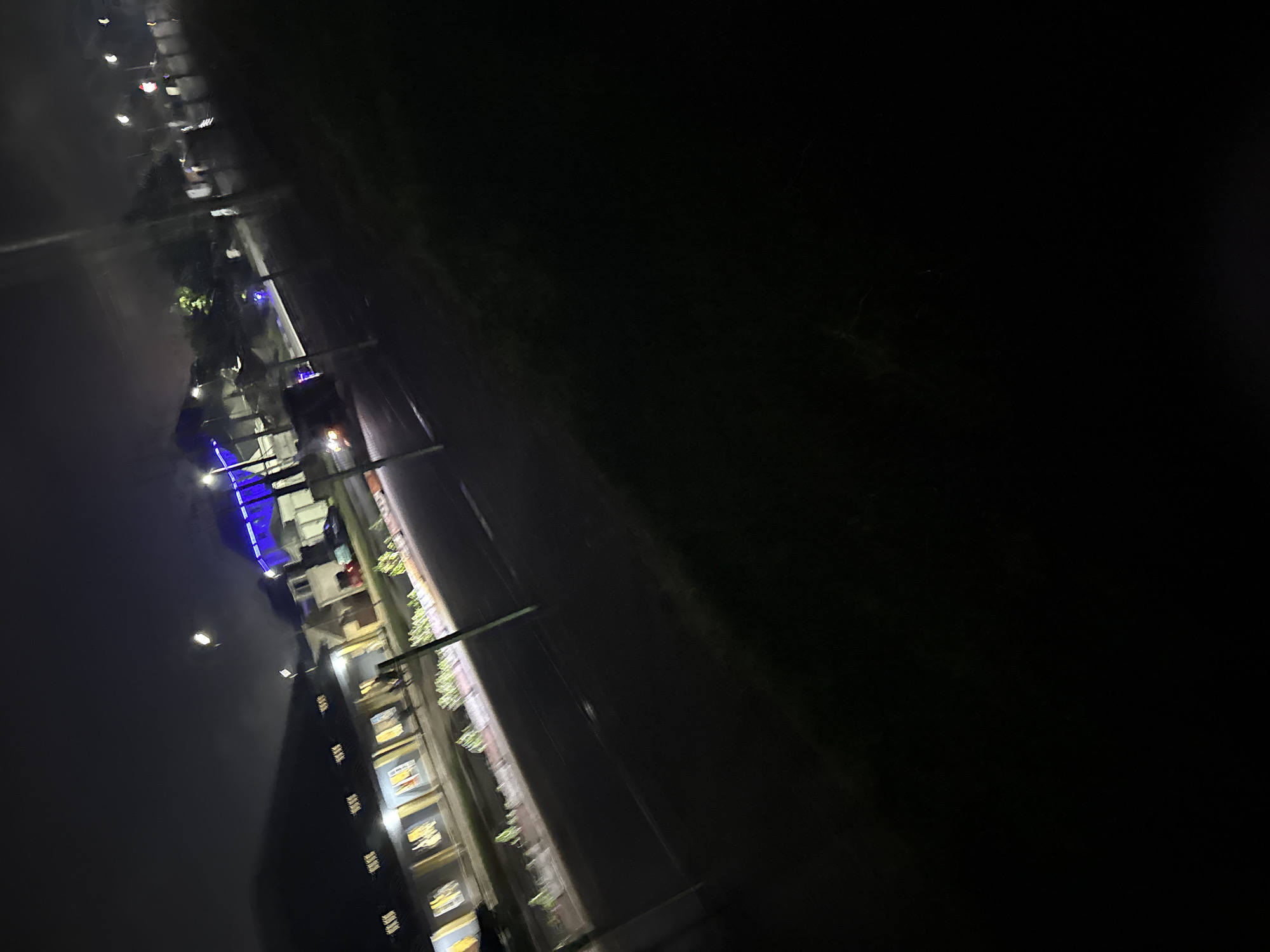 The image depicts a nighttime scene on a street, possibly in Uyo, Nigeria, given the hint. A building with illuminated windows runs along the left side of the frame, appearing to be a school or institution with multiple rooms. Streetlights are visible along the road, casting illumination. In the background, another structure is lit with bright blue lights, possibly a sports facility or event venue. The road appears to be asphalt, with a slight tilt to the right in the composition. The foreground is dominated by dark grass. The overall impression is one of urban infrastructure illuminated by artificial light under a dark sky. There are no identifiable people or specific actions clearly visible in the image, due to the low light conditions and motion blur.