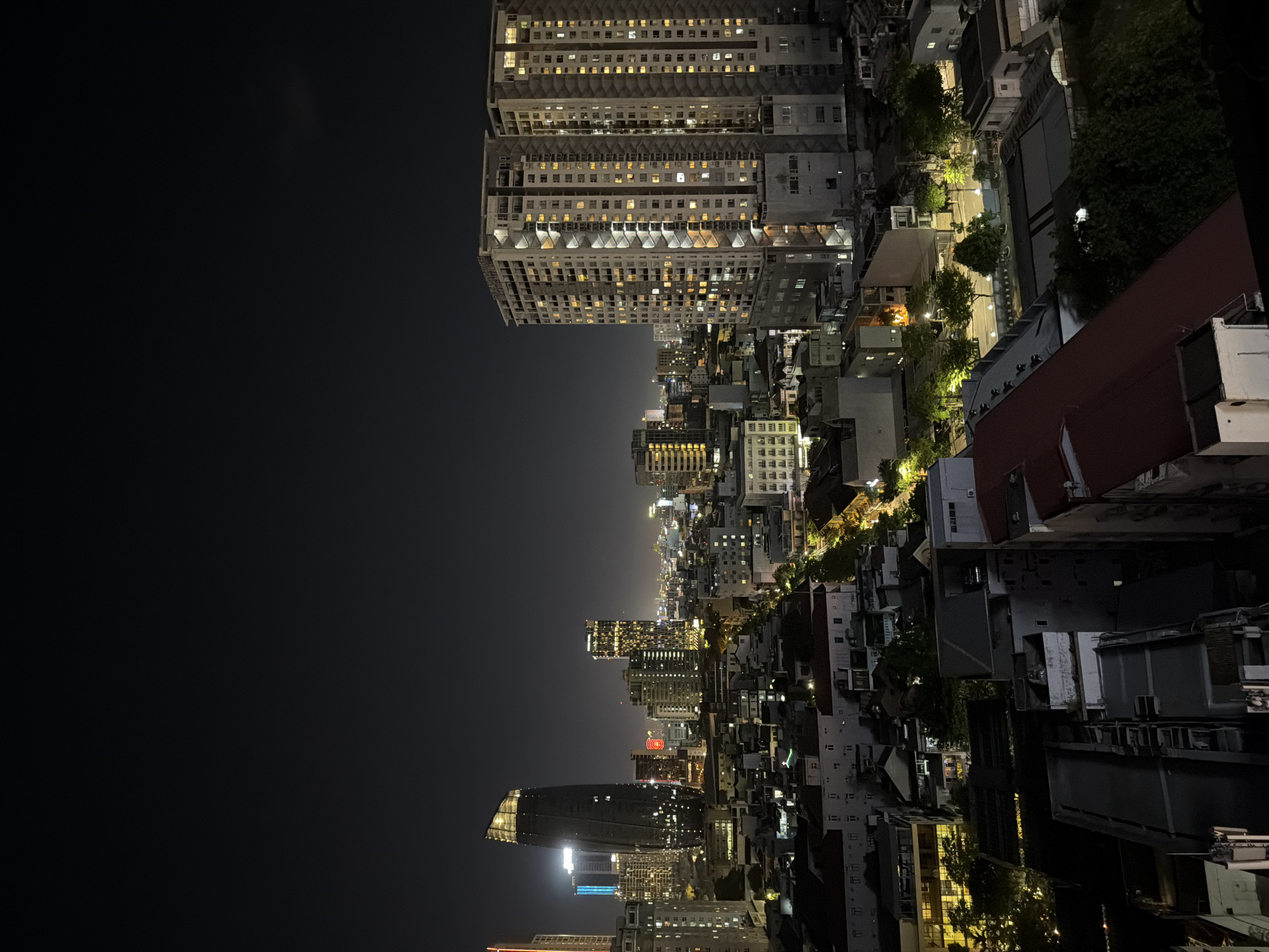This image captures a nighttime aerial view of the city of Đà Nẵng, Vietnam, focusing on its urban landscape and architecture. The foreground is dominated by the rooftops of various buildings, ranging from low-rise structures to more modern residential complexes, some with red tile roofs.
The midground reveals a densely packed cityscape with a mix of building types. A prominent feature is a tree-lined street illuminated by streetlights, casting a warm glow on the surrounding architecture. Several buildings along this street have illuminated windows, suggesting residential or commercial occupancy.
In the background, the skyline is characterized by several tall, modern skyscrapers. One particularly distinctive building, with a curved, metallic facade, stands out with its illuminated upper sections and windows. To its right, two very tall, rectangular apartment buildings are brightly lit, showcasing numerous windows arranged in a grid pattern. These buildings are a focal point of the cityscape.
Further into the distance, the city lights create a hazy glow against the dark night sky, indicating a sprawling urban area. The overall scene is one of a vibrant, active city at night, with a blend of older and newer architectural styles. There are no discernible people or specific actions depicted, the image focuses on the static urban environment. The environment is urban, with clear signs of development and infrastructure.
