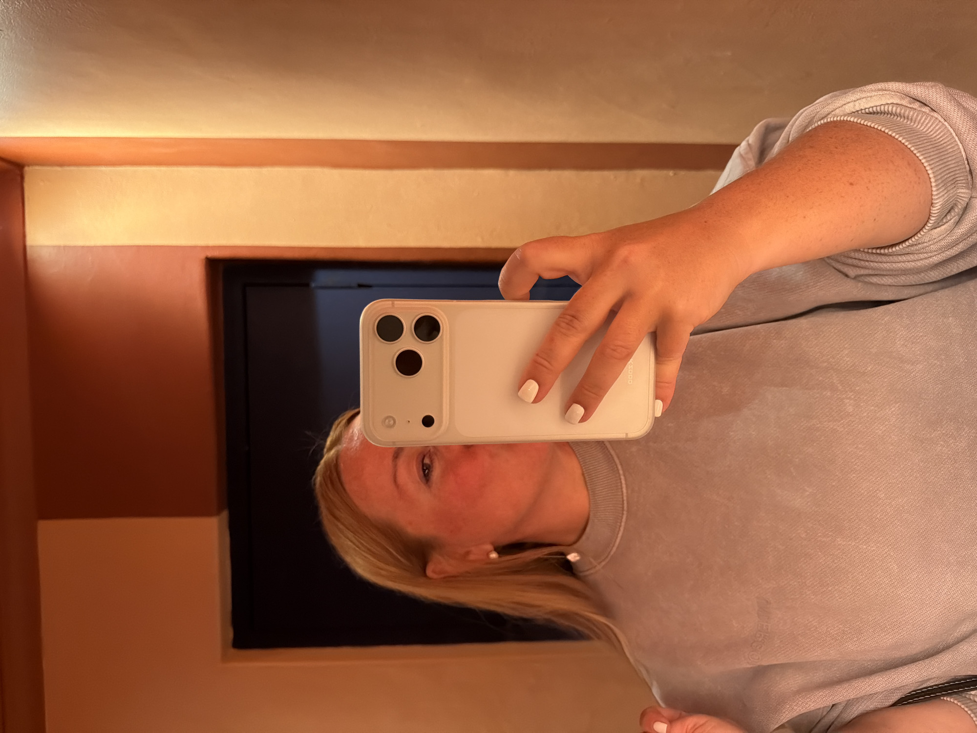 A woman takes a selfie indoors, using a white cell phone with three camera lenses. She has light skin, blond hair, and wears a gray sweatshirt. The background includes a doorway and a wall painted in warm, neutral colors. The location is not discernible from the image, so it is impossible to determine if it is in Cape Town, South Africa, or any other specific location.
