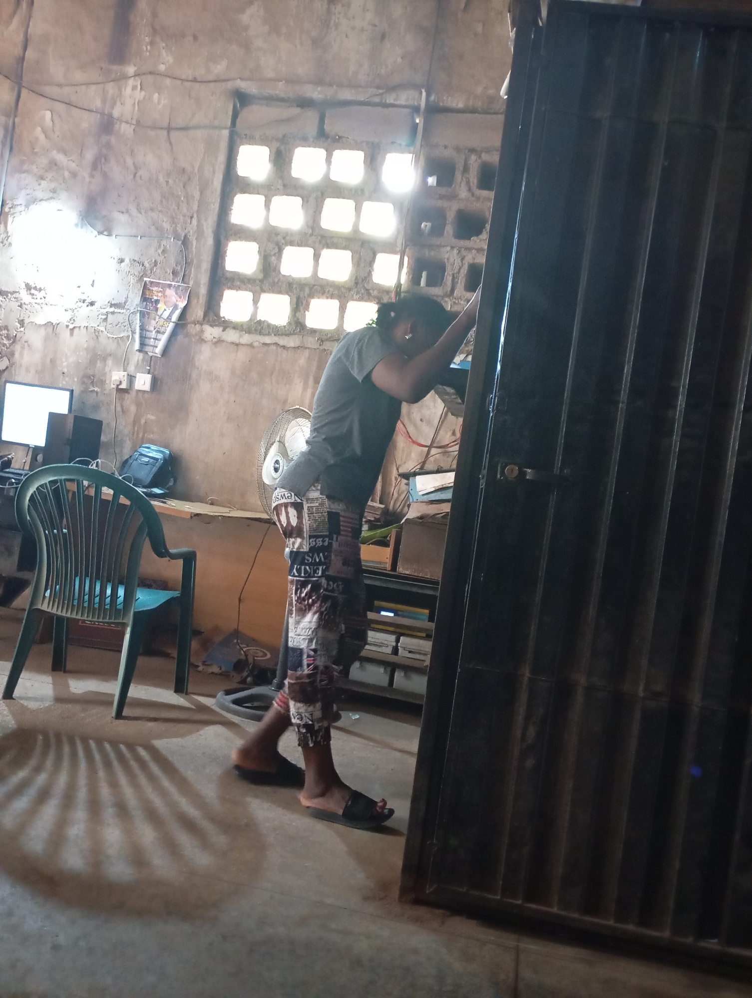 In Shendam, Nigeria, a person stands with their back to the camera, leaning against a large, dark, corrugated metal door or gate. The individual is wearing a grey t-shirt and patterned trousers with text on them. Their feet are clad in black flip-flops. The environment appears to be an indoor space with rough, concrete walls. To the left of the person, a desk holds a computer monitor and a black computer tower. Next to the desk is a green plastic chair. A large fan is positioned behind the person. Sunlight streams in from a window with multiple square openings, casting a striped shadow on the floor. The room is sparsely furnished and has a somewhat dilapidated appearance, with wires visible on the wall and papers attached to it.