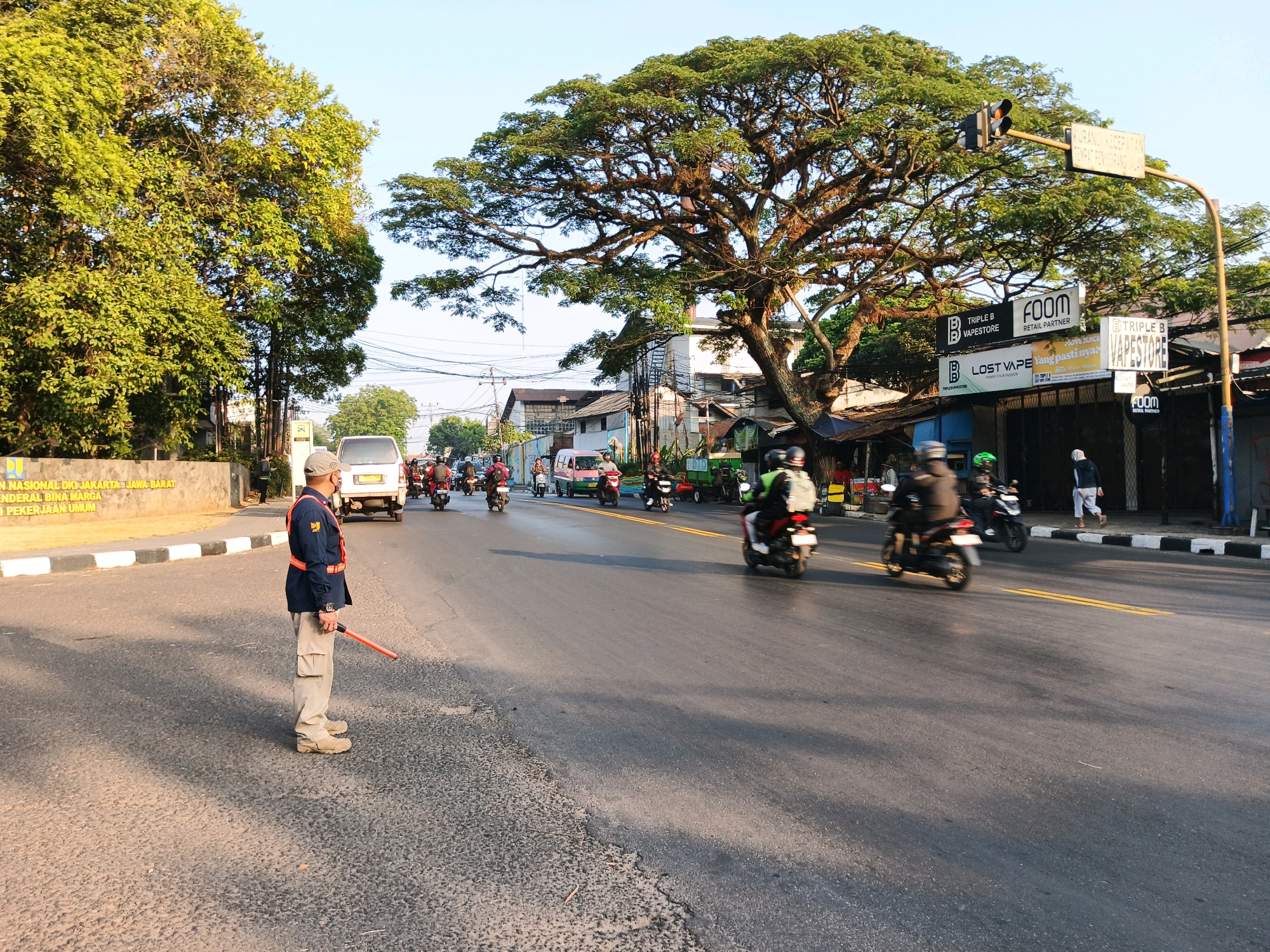 This image depicts a street scene in Bandung City, Indonesia, possibly on a sunny day. A traffic controller stands on the left side of the road, wearing a dark uniform with an orange vest and a cap. He holds an orange baton. Several motorcycles and a white van are on the road, with some of the motorcycles in motion, creating a sense of activity. In the background, buildings with signage are visible, including "FOOM RETAIL PARTNER" and "TRIPLE B VAPESTORE." A large tree with dense foliage is prominent in the mid-ground, casting shadows on the road. The road surface appears to be asphalt, with white and yellow lines marking the lanes. The environment suggests a typical urban street with commercial establishments and moderate traffic.