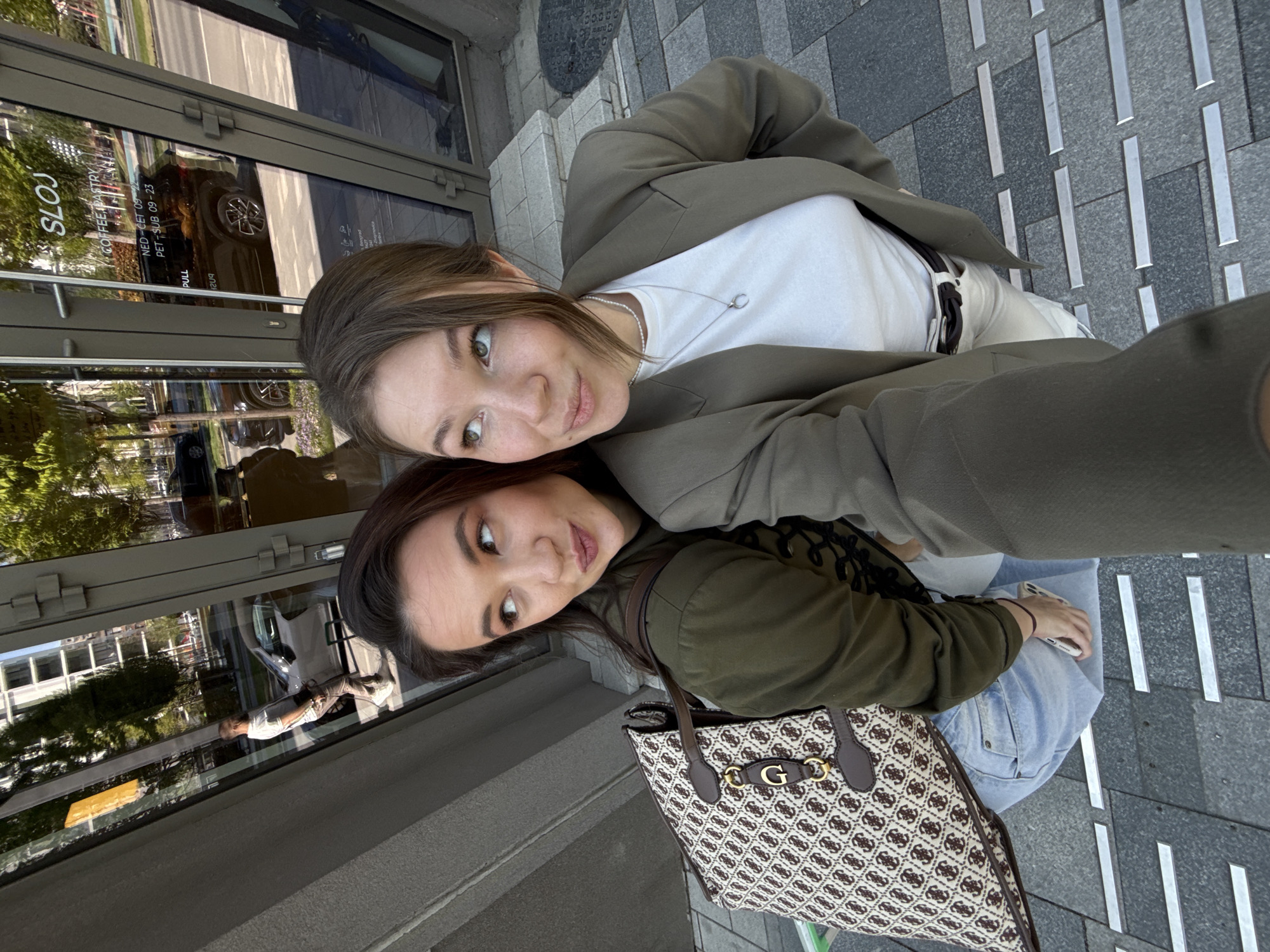 The image features two women taking a close-up selfie outdoors in a city setting, likely Belgrade, Serbia. The woman in the foreground has light brown hair, green eyes, and freckles, and wears a white t-shirt, a silver necklace, and an olive-green blazer. She looks directly at the camera with a slight smile.
Behind and slightly below her, the second woman, holding a white smartphone to capture the selfie, has darker hair and brown eyes, also with a subtle smile. She is dressed in an olive-green jacket and light blue jeans, and carries a patterned handbag featuring a prominent gold 'G' logo, indicating it is likely a Guess brand bag.
They are standing on a paved sidewalk with a mix of grey and light grey patterned tiles, interspersed with reflective metallic strips, possibly tactile paving. Behind them is a large glass window, reflecting the urban environment. These reflections show several vehicles, including a dark car and a light-colored van, as well as trees and parts of other buildings. A distant figure is also visible in the window's reflection.
The scene is brightly lit, suggesting daytime with clear weather. Visible text reflected on the glass window, partially reversed, includes "SLOJ", "COFFEE PASTRY", and what appear to be opening hours: "NED - CET 09 - 23" and "PET - SUB 09 - 23". These likely translate to "Sunday - Thursday 9 AM - 11 PM" and "Friday - Saturday 9 AM - 11 PM" (09:00-23:00). The word "PULL" is also faintly visible on a door or window pane. The overall atmosphere is casual and friendly.