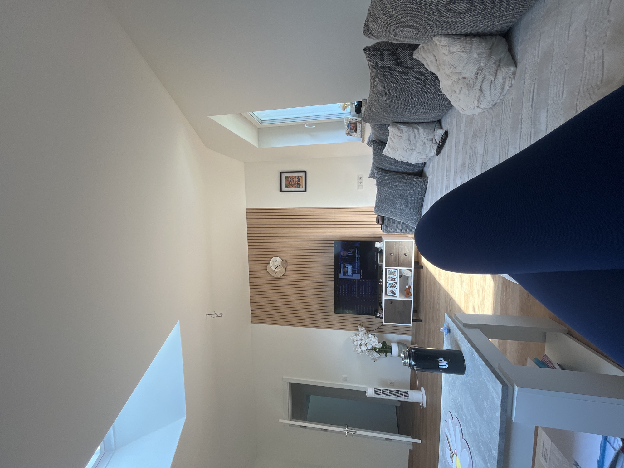 The image depicts an indoor living room scene bathed in natural light from a skylight and a window. The room has white walls and light-colored wooden flooring. A large sectional sofa with numerous grey and white patterned cushions occupies the right side of the frame. A remote control is visible on the sofa. In the center of the room, a modern entertainment center with a dark wood and white finish stands against a wall paneled with light wood slats. A television screen is displaying content. To the left of the entertainment center, a white orchid in a pot is placed on the floor. Further left, a white oscillating tower fan stands near an open door leading to another room. A black thermos with the word "UP" printed on it in white sits on a white and grey marble-topped coffee table. A person's legs, clad in dark blue leggings, are visible in the foreground, suggesting they are seated on the sofa or near the coffee table. The environment suggests a modern, well-lit, and comfortable living space. There is no indication of the location being Garmisch-Partenkirchen, Germany.