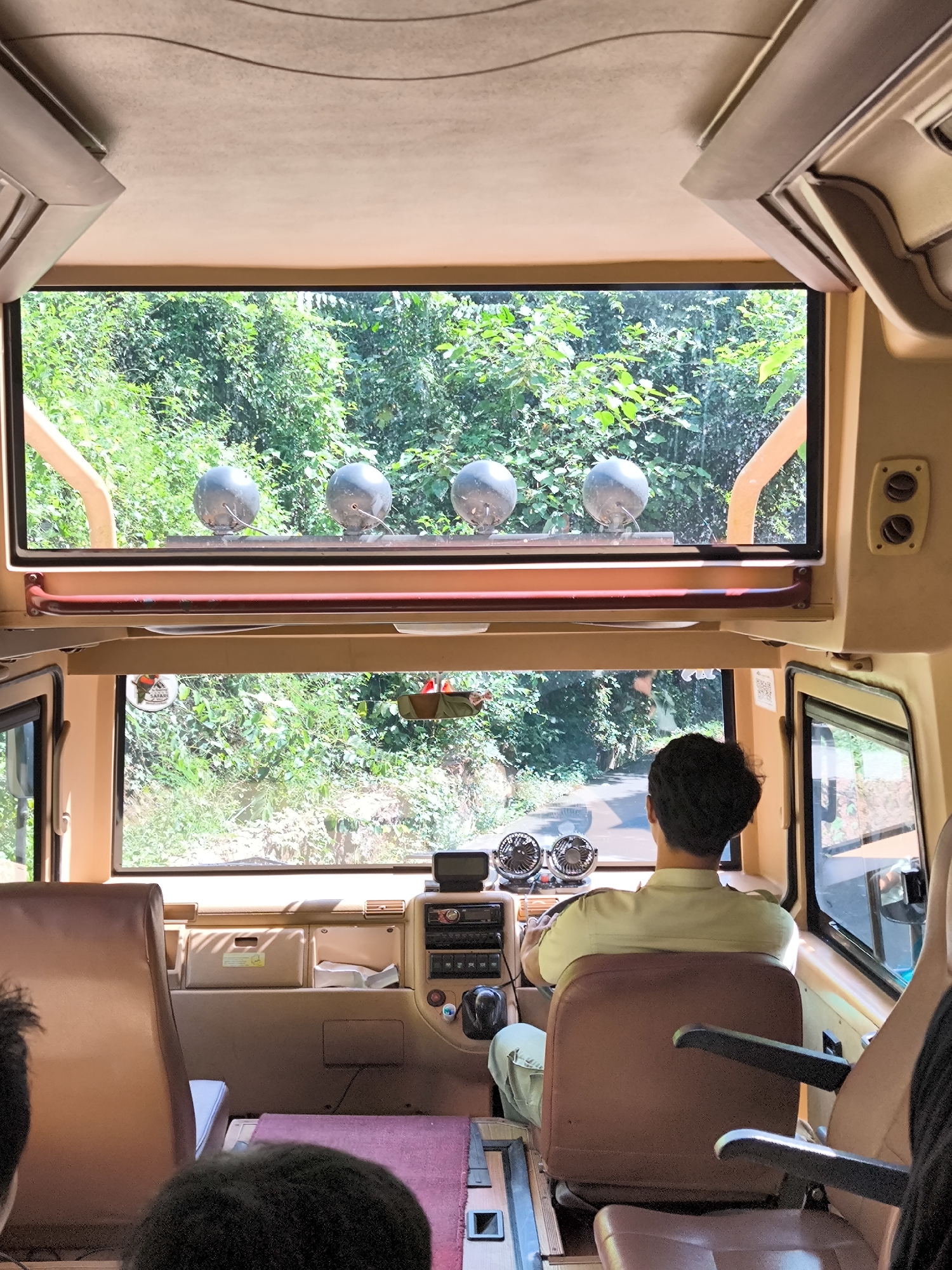 A view from inside a bus looking forward, with the driver in the foreground, facing away from the camera and holding the steering wheel. The driver is wearing a light-colored collared shirt and light-colored pants. The bus has tan upholstery and a tan headliner. The dashboard is visible, with various controls and two circular fans. Through the windshield, a lush green forest is visible, with a road winding through it. Above the windshield, there are four spherical spotlights mounted on the exterior of the bus. The image appears to be taken during the daytime.