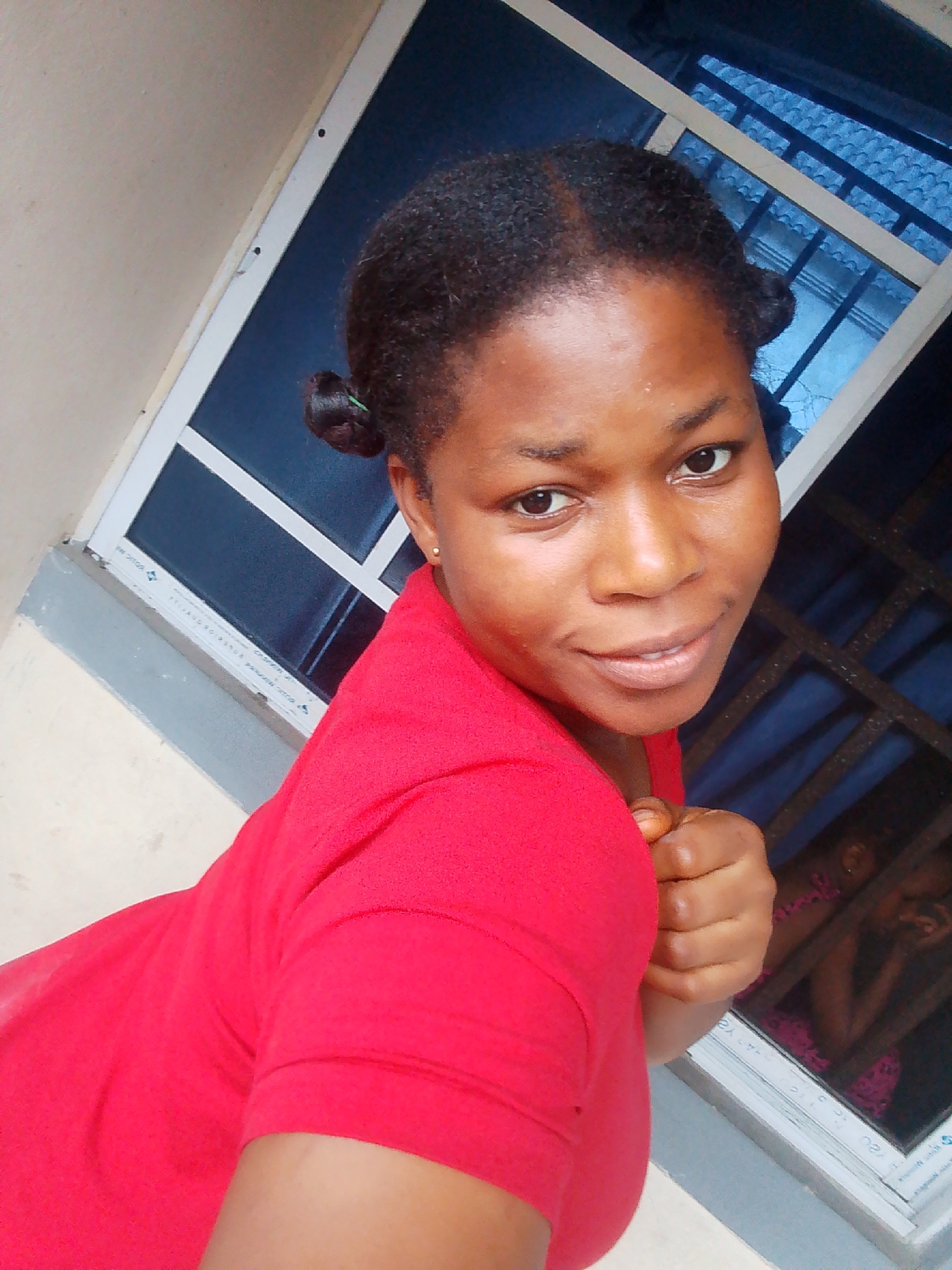 The image shows a young woman posing for a selfie. She has dark skin and her hair is styled in two buns. She is wearing a red top and her left arm is bent, with her fist clenched. She is looking at the camera with a slight smile. In the background, there is a window with blue bars and a glimpse of another person or people behind it, possibly children wearing pink. The setting appears to be outdoors, with a wall and window frame visible. The lighting suggests it is daytime. There is no visible text in the image. The location is identified as Yenegoa, Nigeria.
