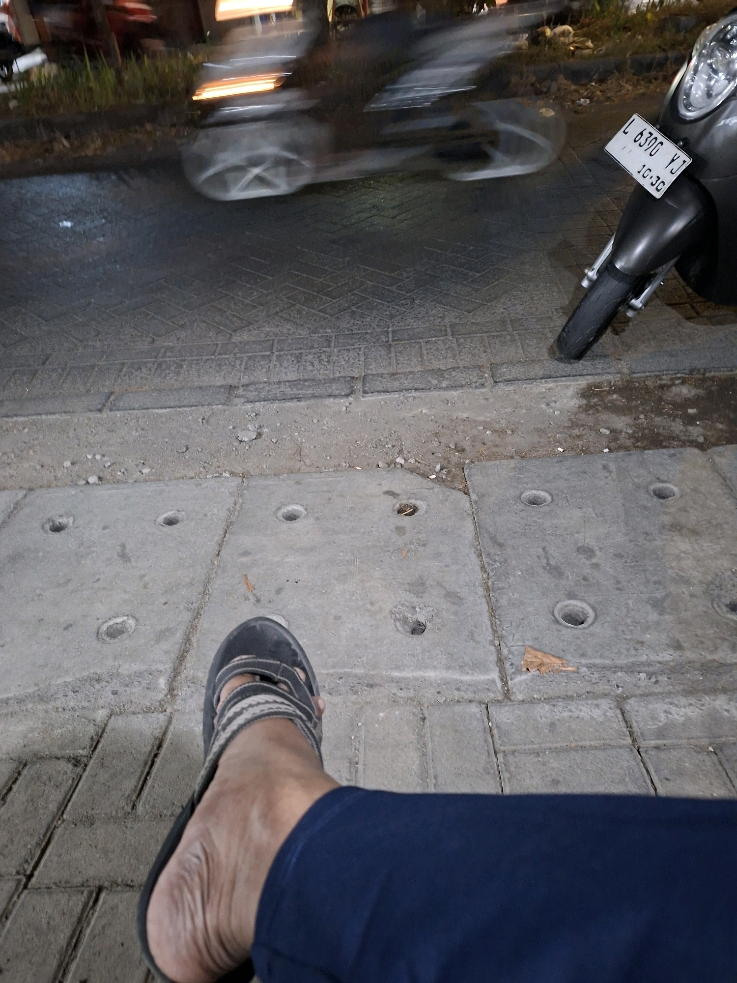 The image depicts a low-angle view of a person's leg and foot wearing a dark sandal and navy blue pants, resting on a concrete sidewalk. In the background, a motorcycle with a license plate reading "L 6396 YJ" is parked. Further in the background, a blurred image suggests a busy street scene with moving vehicles, indicated by streaks of light and motion blur. The environment appears to be a street in Surabaya, Indonesia, characterized by the concrete pavement, storm drains, and urban elements. The lighting suggests it is evening or night.