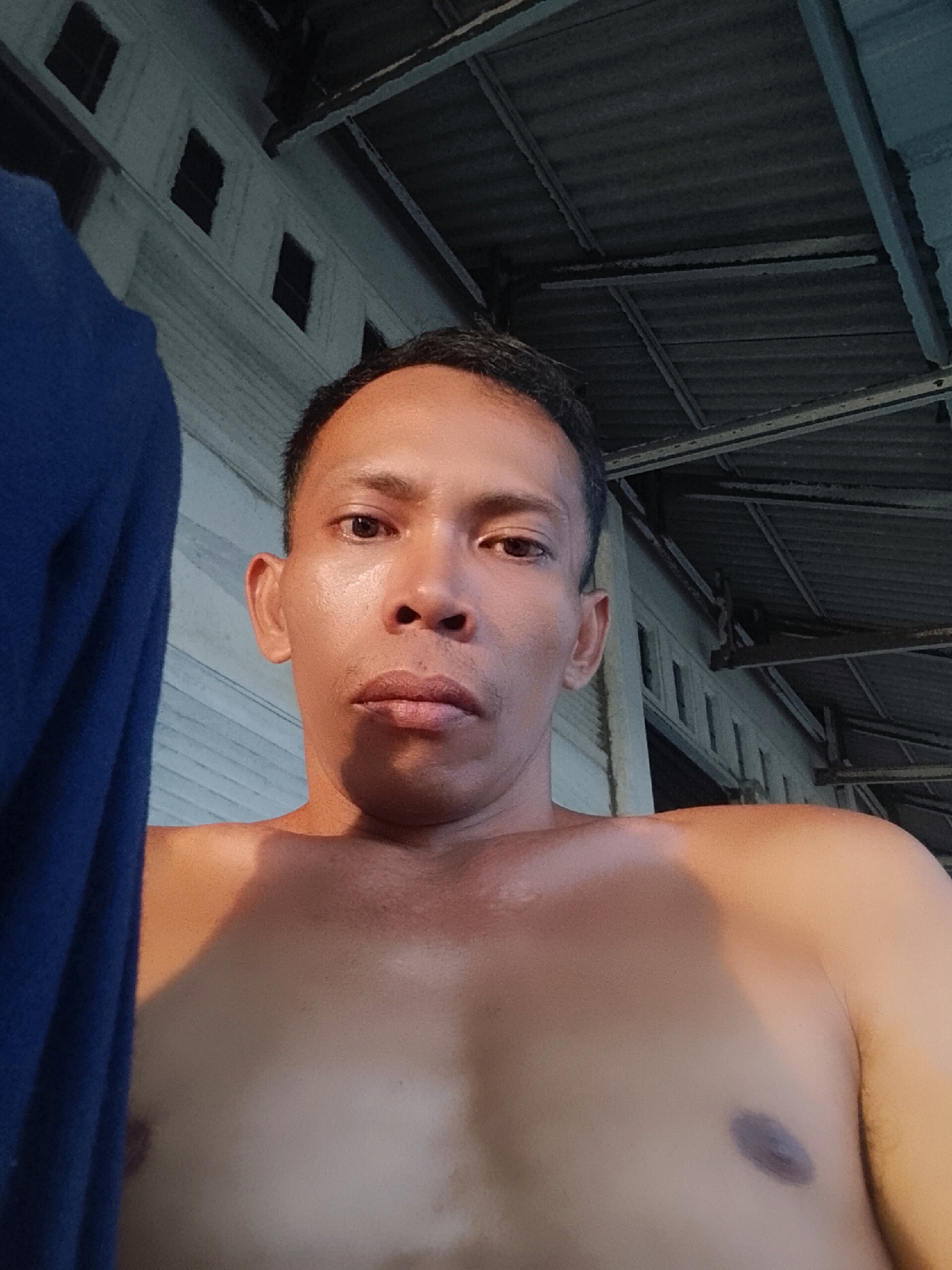 A shirtless man is posing for a selfie. He has short dark hair, brown eyes, and is looking directly at the camera with a neutral expression. He is standing outside, possibly on a porch, as evidenced by the corrugated metal roof and white wall with small square windows visible in the background. A blue fabric drapes to the left of the frame. The location is possibly Bukit Ketok, Indonesia.