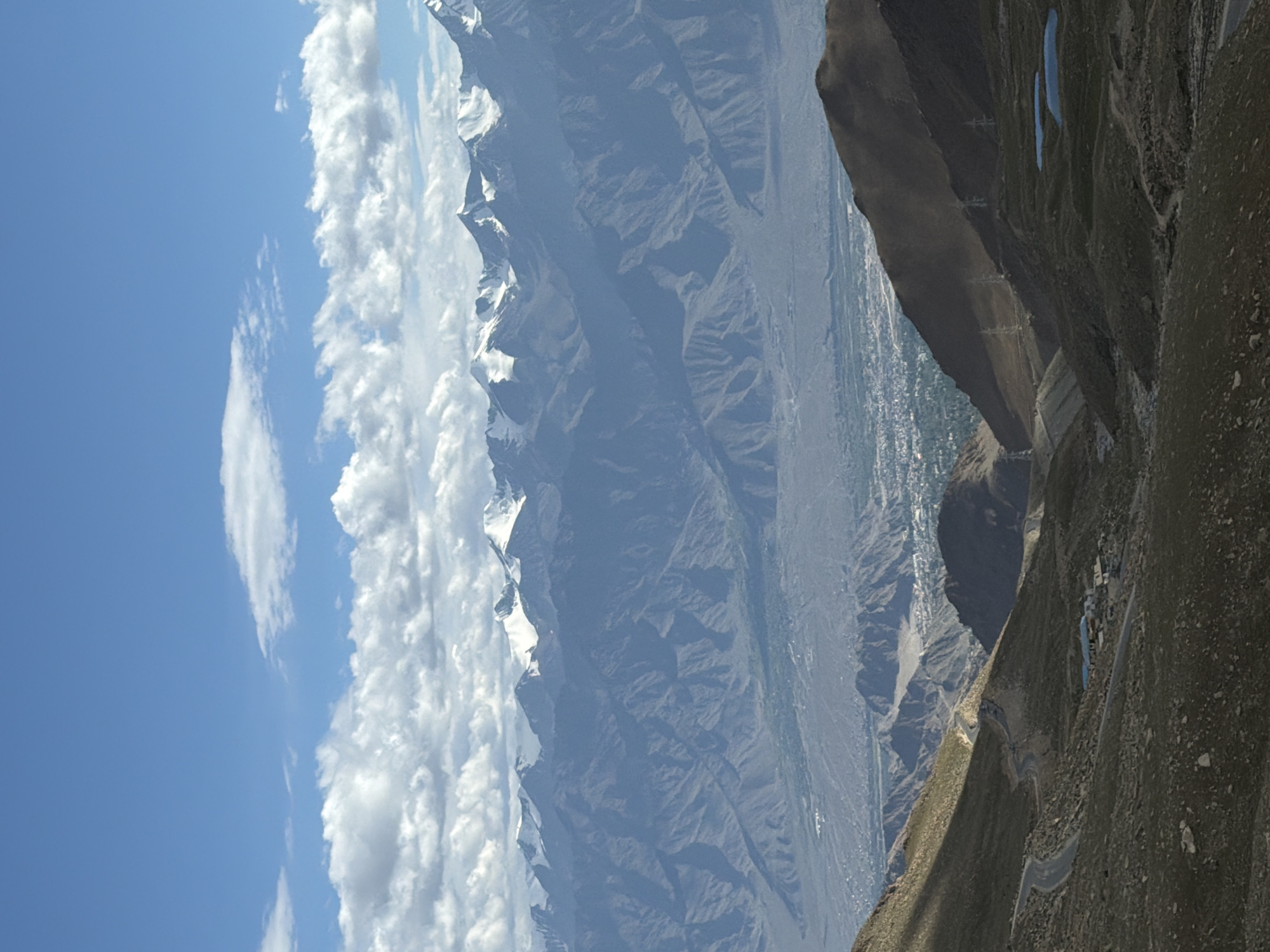 This is a wide-angle vertical photograph of a mountainous landscape. In the foreground, a winding road descends a rocky, sparsely vegetated slope. A cluster of buildings with blue roofs is visible near the road. Further down the slope, a valley opens up, revealing a town with many buildings. Beyond the valley, a range of large, rugged mountains stretches across the frame, many of their peaks covered in snow. The sky above is blue with scattered white clouds. This scene is consistent with views from the Khardung La Pass, a high mountain pass in the Ladakh region of India, near the city of Leh.