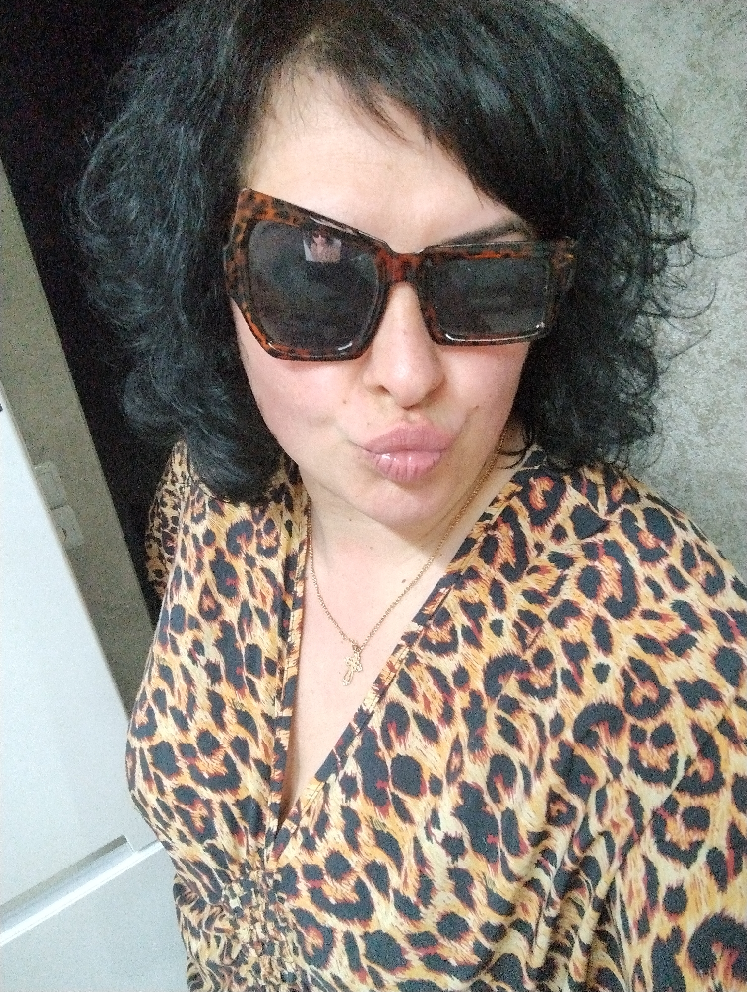A woman is taking a selfie indoors. She is wearing a leopard-print top and large, dark sunglasses. She has dark, curly hair and is wearing a gold necklace with a cross pendant. Her lips are puckered. The background includes a dark doorway or wall and a light-colored wall. There is no visible evidence to suggest that the image was taken in Grishanki, Russia.