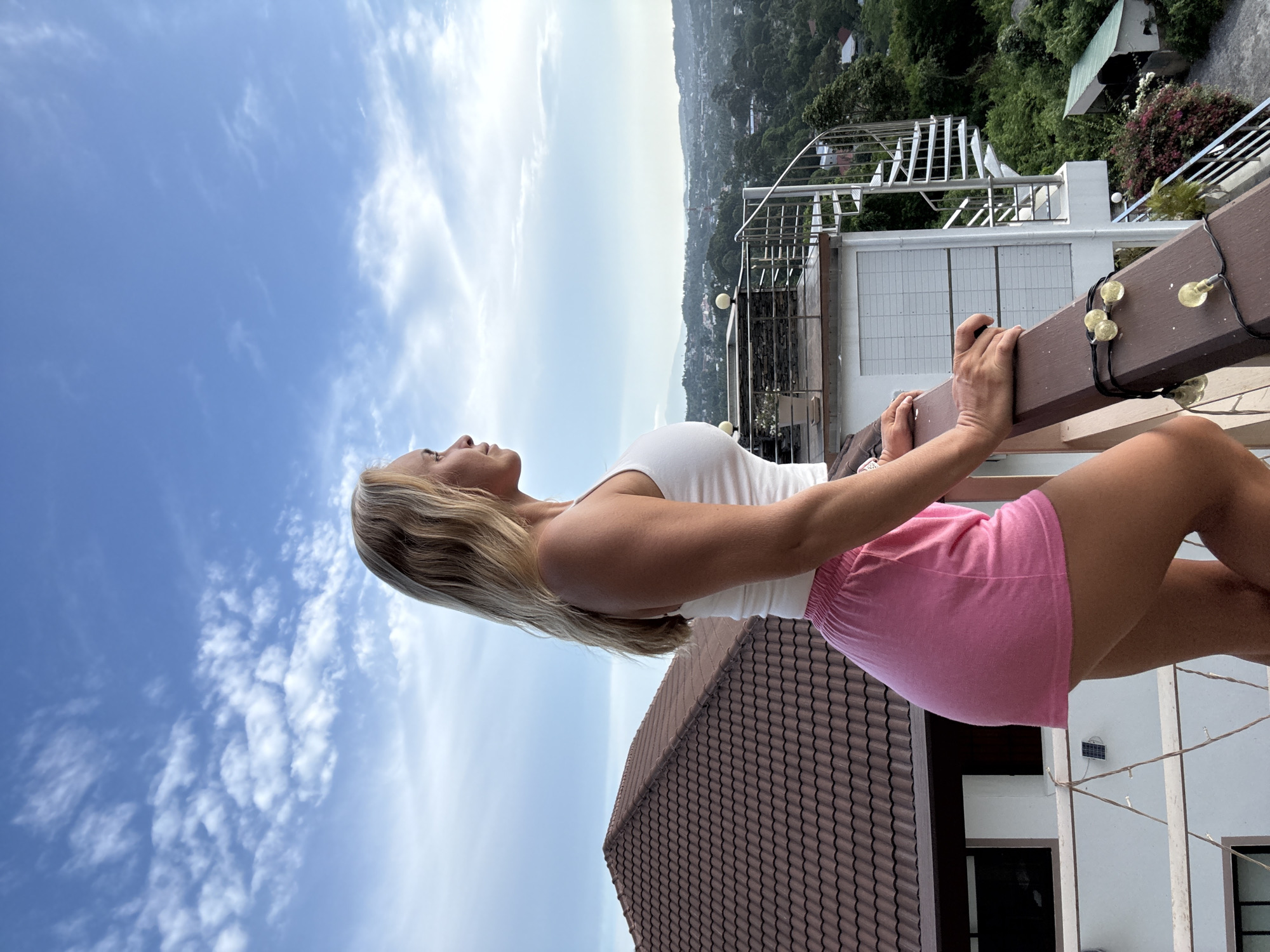 A blonde woman stands on a balcony overlooking a city in Ko Samui District, Thailand. She is wearing a white tank top and pink shorts and is looking out at the view. The balcony has a brown railing with string lights. In the background, there is a tiled roof of a house, a spiral staircase, and lush greenery. The sky is blue with scattered clouds.