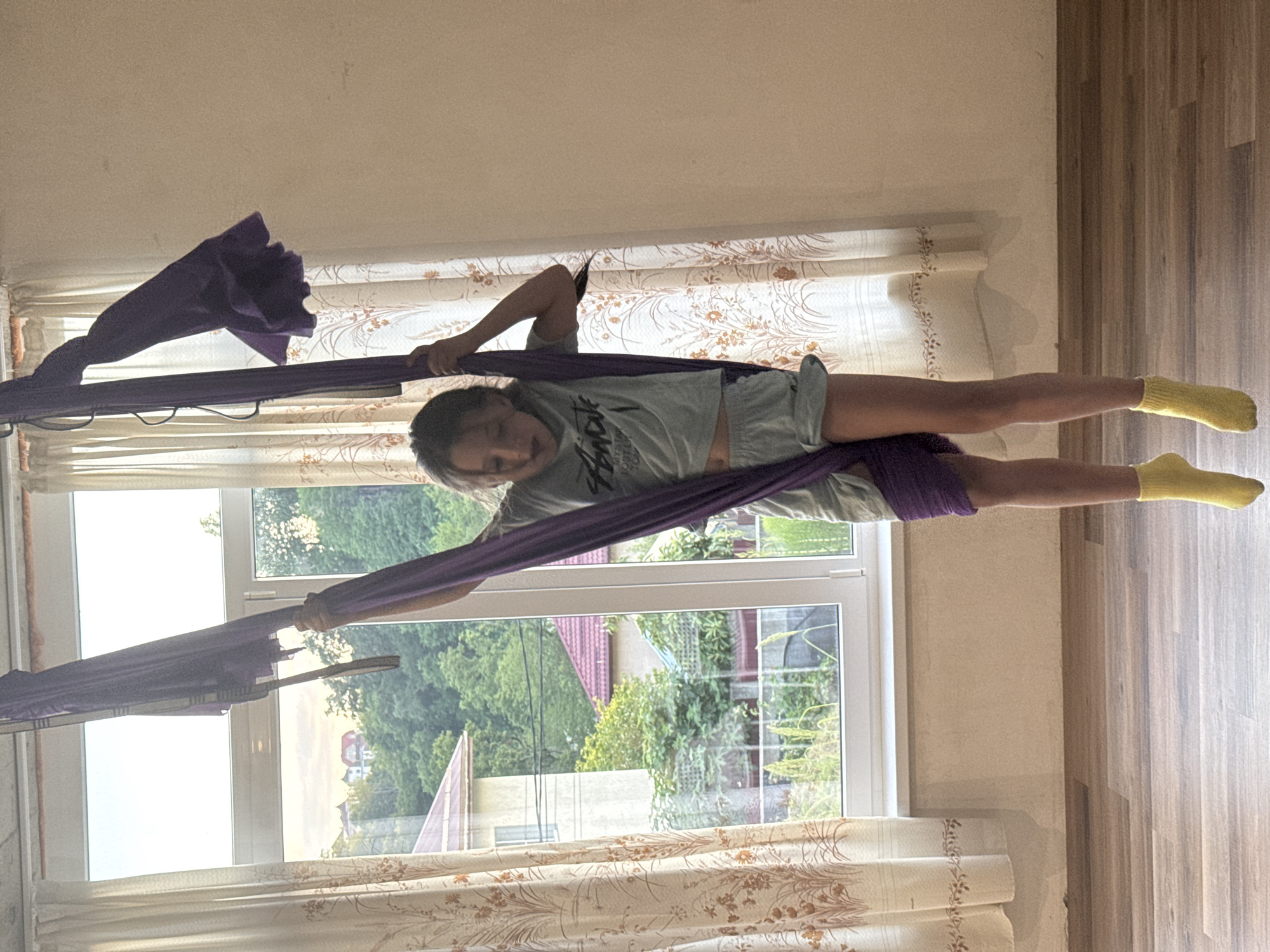 A young person is performing aerial yoga on a purple fabric hammock indoors. The hammock is suspended from above, and the individual is stretched out horizontally, with their legs extended and one hand gripping the fabric for support. They are wearing a light-colored t-shirt, shorts, and yellow socks. The background features a window with a view of trees and houses, suggesting a residential setting, likely in Verkhniy Yurt, Russia. Sunlight is coming through the window, casting shadows on the floor and illuminating the room. The curtains have a floral pattern. The overall mood appears to be one of focus and physical activity. There is visible text on the t-shirt, but it is not clearly legible.