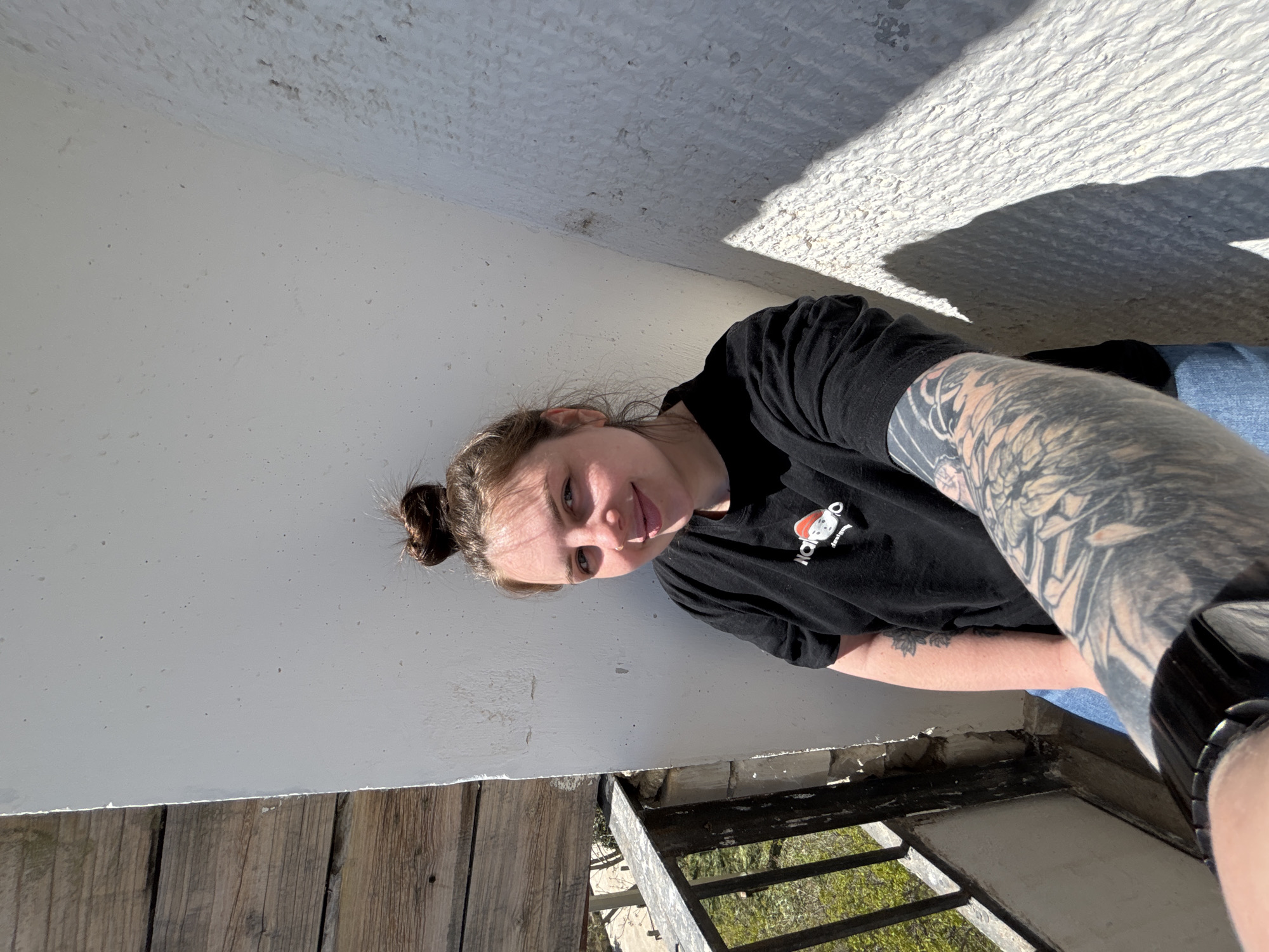 A woman is posing for a photograph, appearing to be outside in what seems to be a partly shaded, outdoor area. The background includes what appears to be a white wall or structure, and below it there's a wooden surface or platform, alongside a metal railing and green vegetation visible through the railing. The woman is wearing a black T-shirt and has a tattoo covering most of her visible arm. There is no explicit visual evidence indicating that this picture was taken in Chornomorskoe, Ukraine.