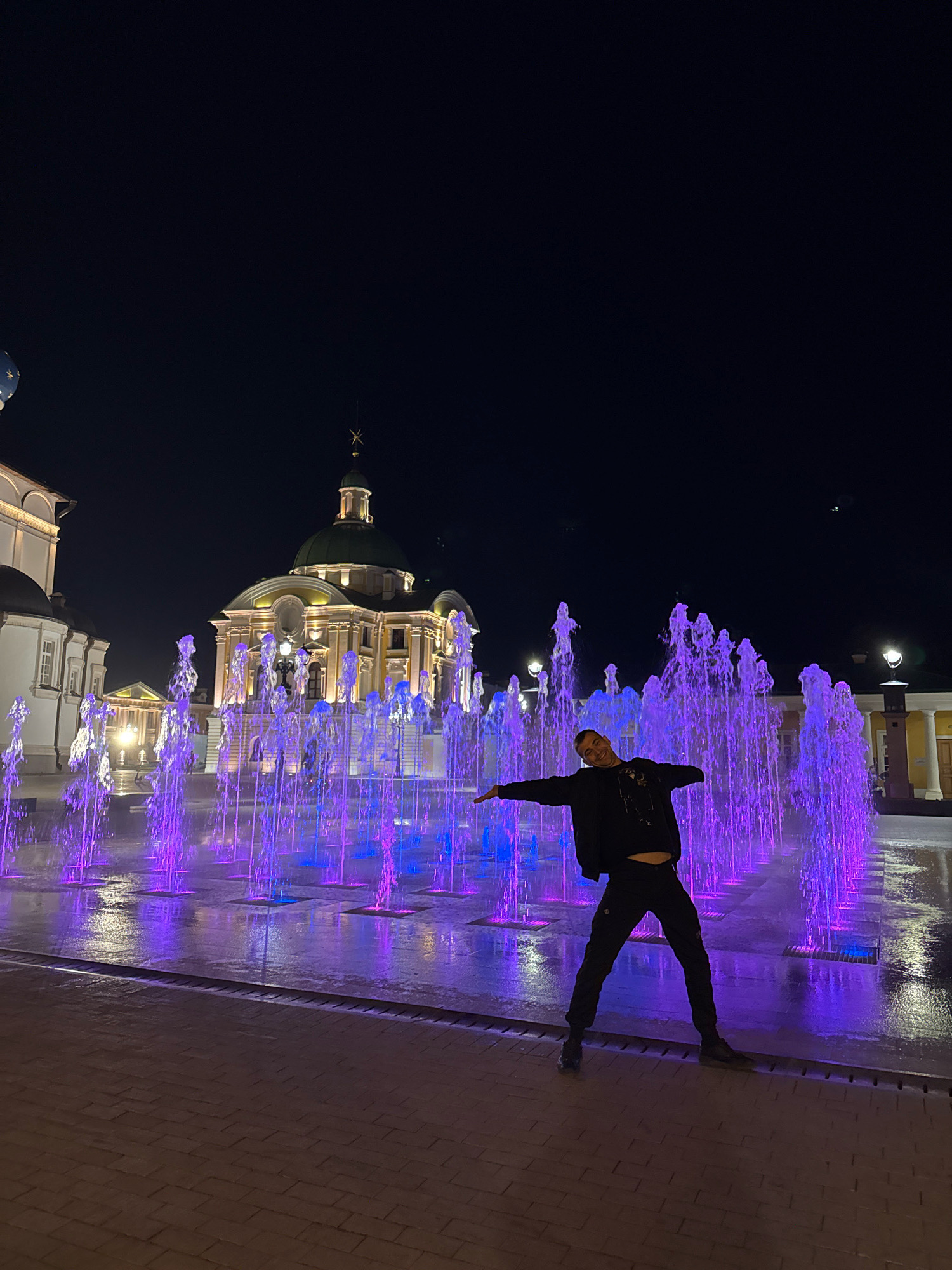 A man stands with his arms outstretched in front of a fountain illuminated with purple lights at night. The fountain is in a plaza with a brick pavement. In the background, a building with a green dome is visible, along with other buildings with ornate architecture. The scene appears to be in the city of Tver, Russia.