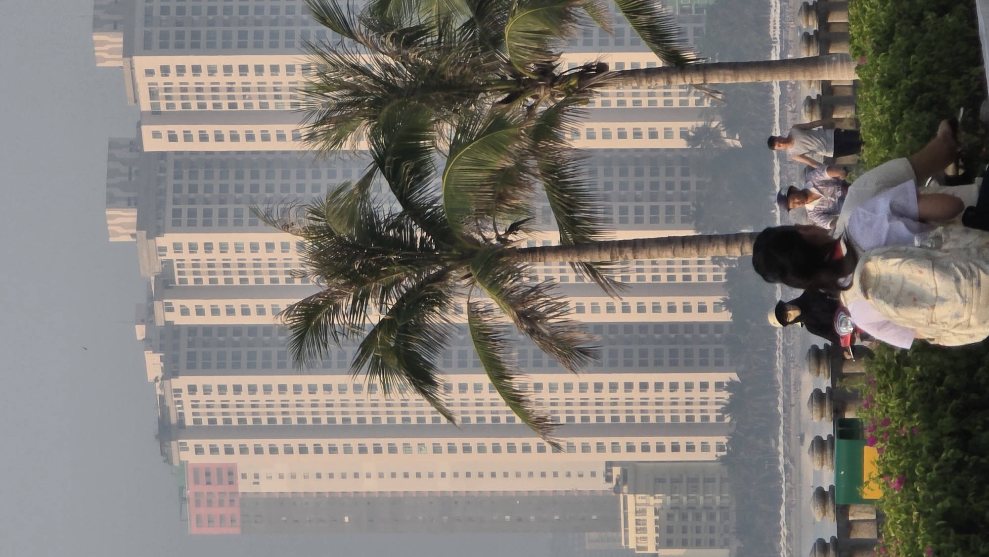 The image depicts a park scene in Bắc Nha Trang, Vietnam, with tall apartment buildings in the background. Two palm trees are prominent in the foreground. Several people are visible along a waterfront promenade. One person is seated, and others are walking along the pathway. The environment appears hazy, with the buildings receding into the distance. The promenade is bordered by a low wall and greenery.