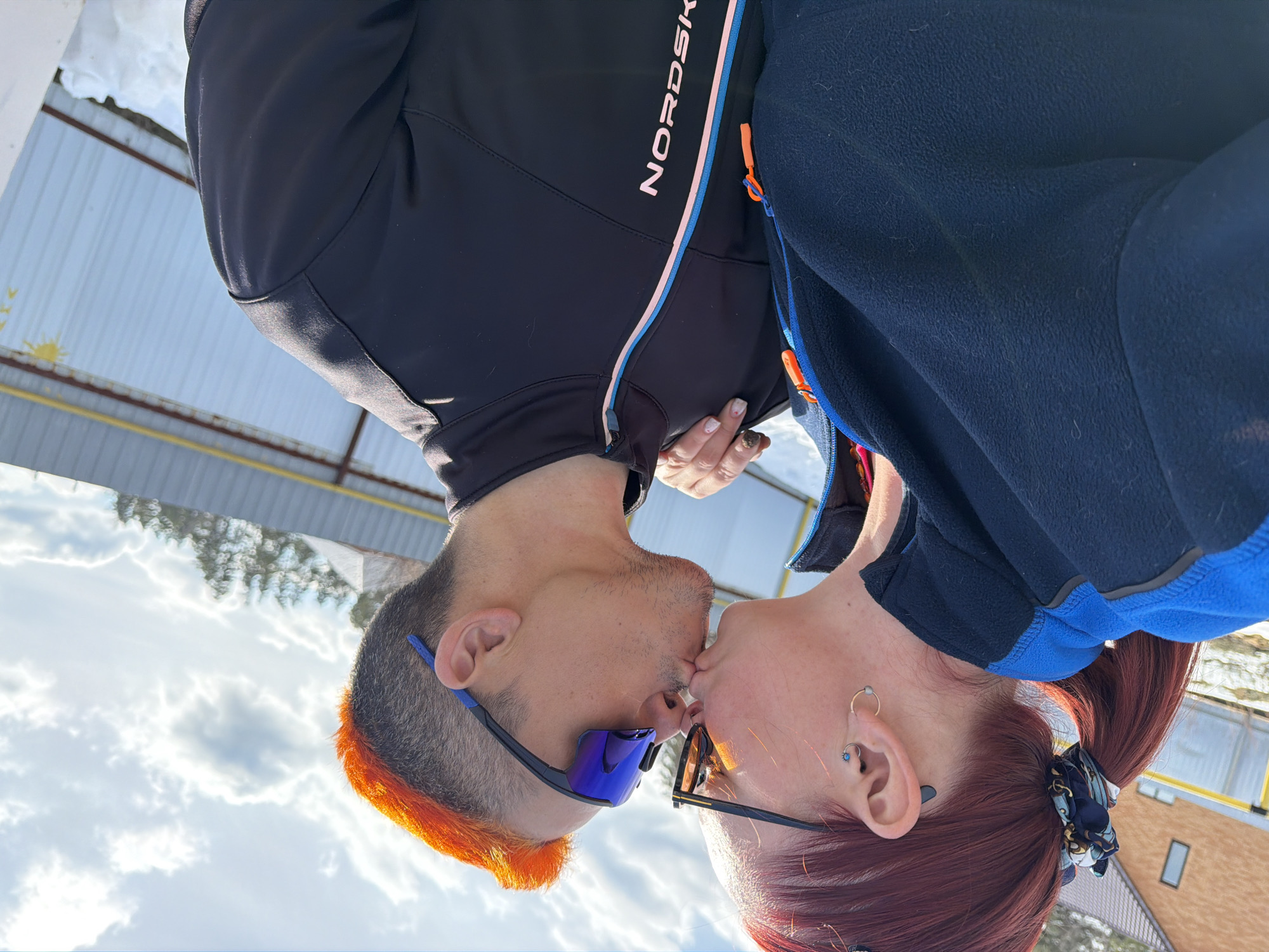 Two people are kissing. The person on the left has dark hair with orange tips and is wearing blue sunglasses and a black jacket with white and pink accents. The person on the right has red hair pulled back in a ponytail and is wearing black glasses and a dark blue fleece jacket with blue accents. The photo appears to be taken outside in a snowy environment under a slightly cloudy sky. The location may be Chaikovsky, Russia, based on the prompt's request. Visible in the background is a metal roof structure, trees, and a building with a window.