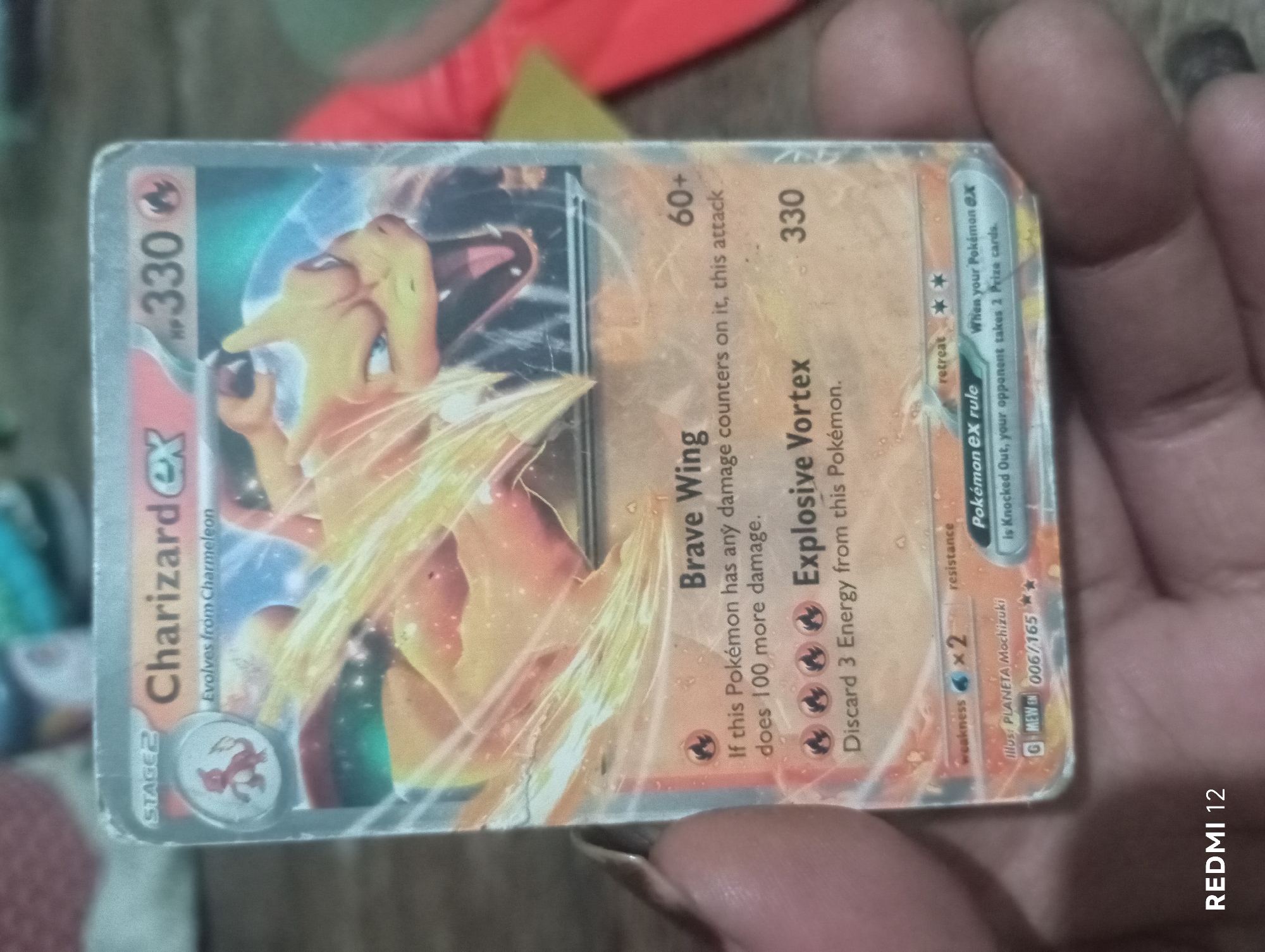 A close-up, overhead shot shows a hand holding a Pokémon trading card. The card is a "Charizard EX" and is in the "Stage 2" evolution. The card features an illustration of Charizard, a dragon-like creature, breathing fire. The card has text indicating its abilities and stats. The background is blurred but appears to be a wooden surface. The image is likely taken indoors, possibly in a home or a shop. There is no discernible information in the image to suggest it is located in Pagar Alam, Indonesia.
