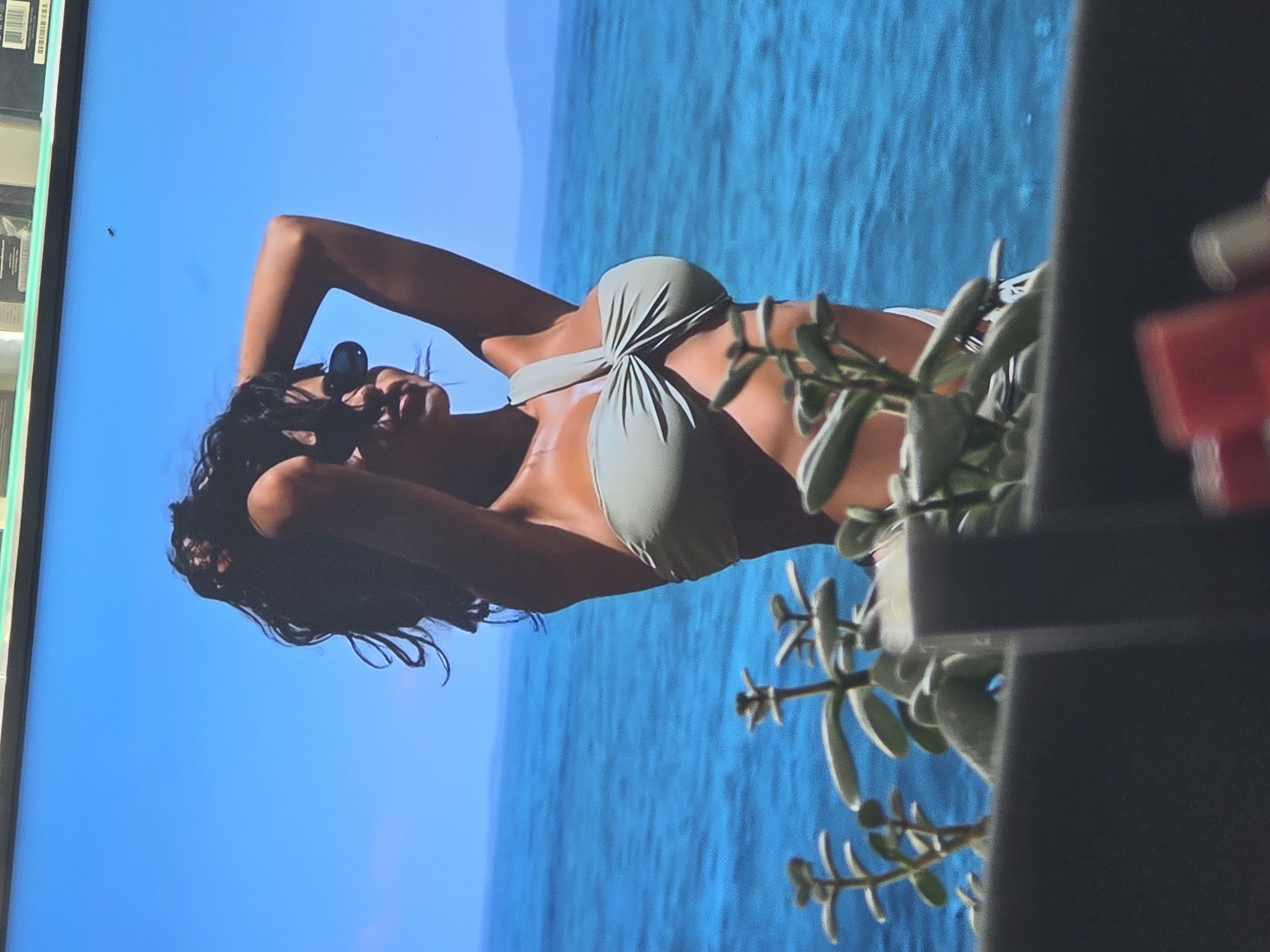 The image shows a woman wearing a light green bikini, posing with her arms behind her head. She is lying down on what appears to be a beach, with the bright blue ocean in the background. The sun is shining brightly, casting a warm glow on her skin. Her hair is dark and wavy, and she is wearing sunglasses, obscuring her eyes. In the foreground, a green plant with thick, succulent-like leaves is partially obscuring the view of the woman. The scene evokes a feeling of relaxation and vacation. The image is taken from a slightly elevated angle, looking down at the woman. There is no visible text in the image. The location context of Varna, Bulgaria, suggests this could be a seaside resort or a beach town. The clear blue sky and bright sunlight indicate a warm, sunny day, possibly during the summer months.