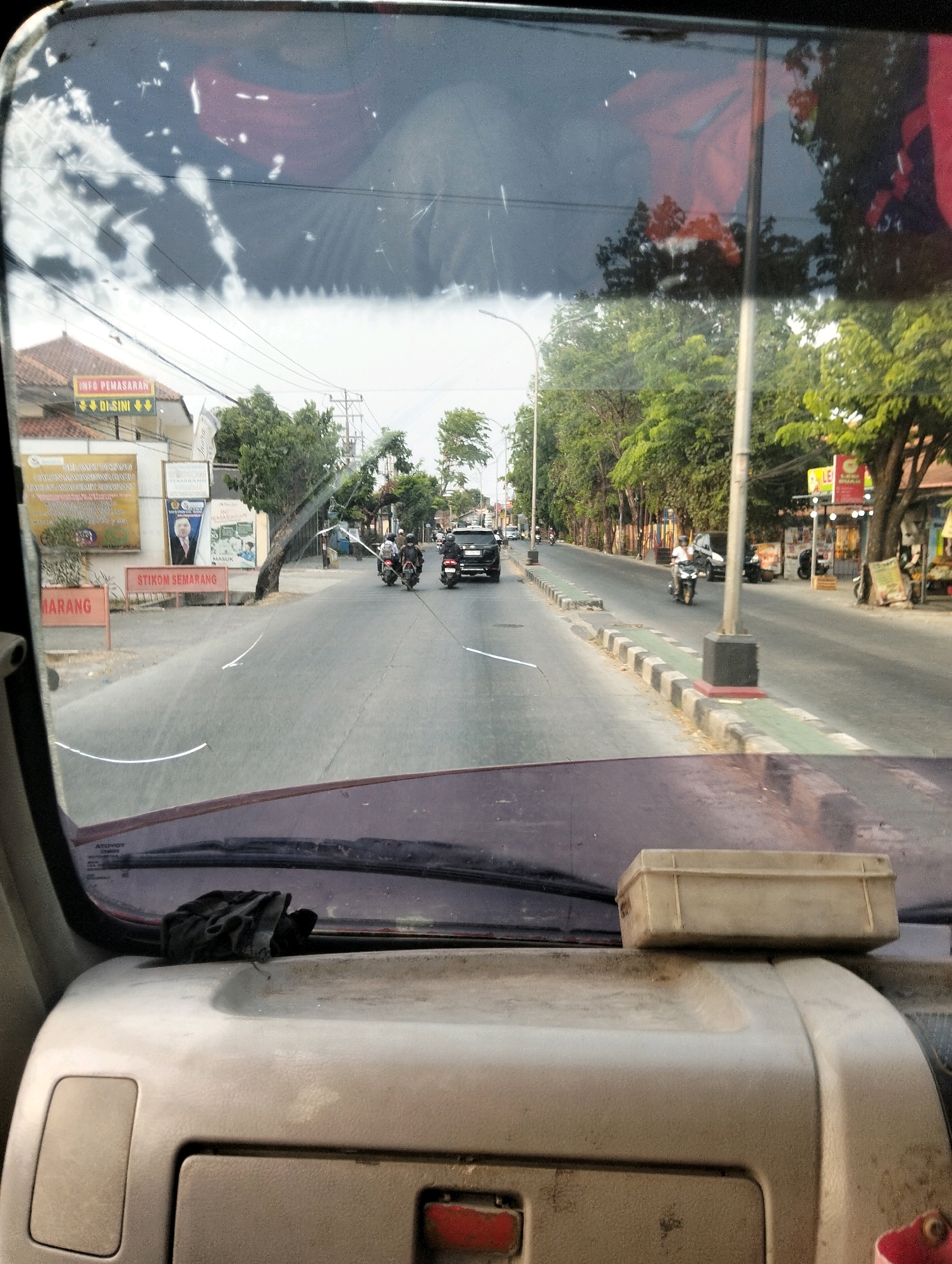 The image captures a view from inside a vehicle looking through the windshield at a street scene in Semarang, Indonesia. The street is paved and lined with trees on both sides. Several vehicles are visible on the road, including motorcycles and a black car. On the left side of the road, there are buildings with signs, one of which appears to be an advertisement for "INFO PEMASARAN" and "DI SINI". Another sign reads "STIKOM SEMARANG". People are seen riding motorcycles, some wearing helmets. The sky is visible through the windshield, reflecting the surroundings. A black cloth is placed on the dashboard of the vehicle, and a light-colored rectangular object is also on the dashboard. The overall impression is a typical urban street scene during the daytime.