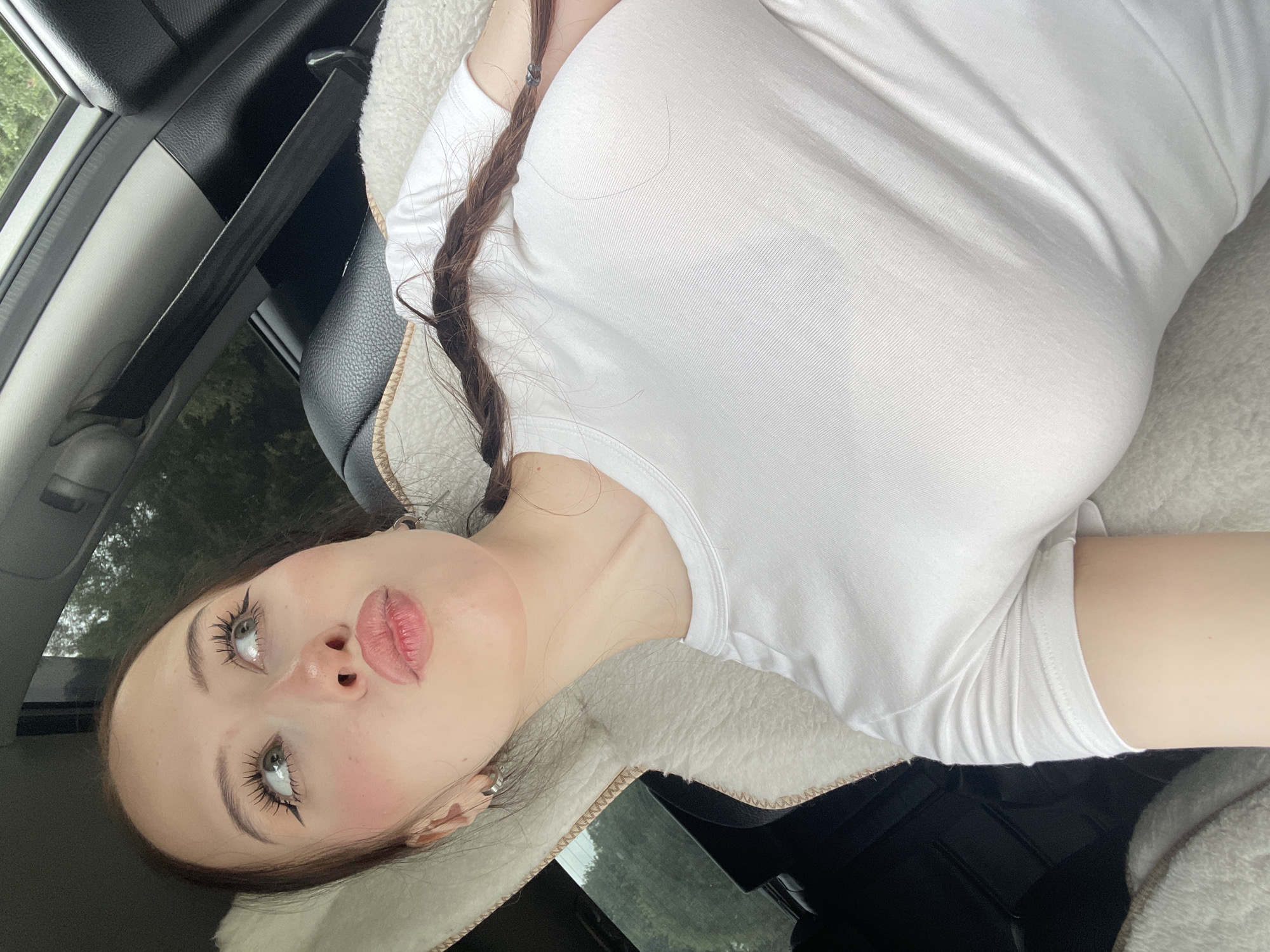 A young woman with long brown hair in a braid, wearing a white t-shirt and hoop earrings, is captured in a close-up selfie. She is sitting in the passenger seat of a car, leaning her head against a white fluffy pillow. Her eyes are looking upwards and to the left, her lips are puckered, and she has noticeable eyeliner and blush. The background shows a car interior with a black seat and a seatbelt, and a glimpse of trees outside the window. The scene takes place in Gavrilovka, Russia.